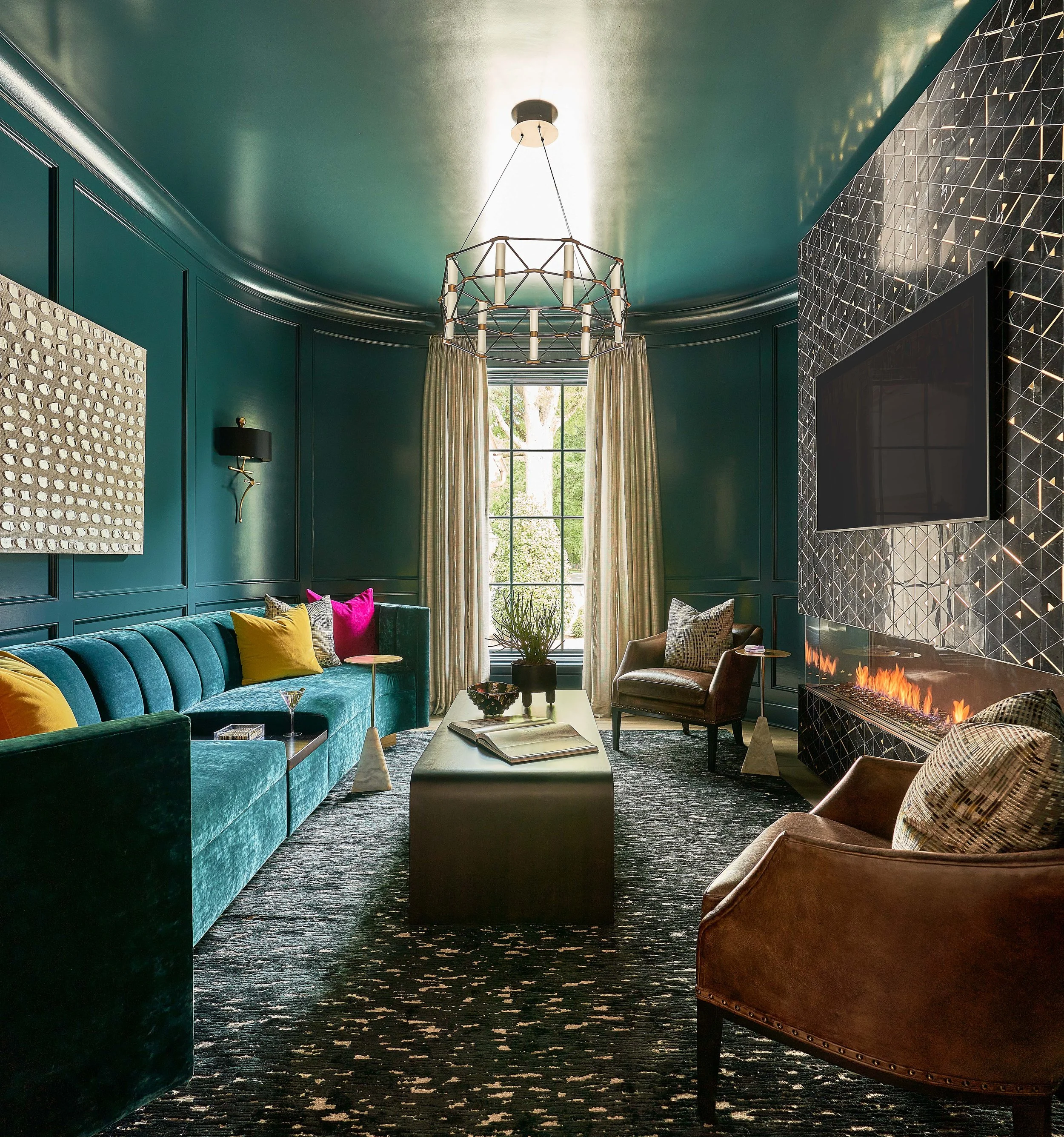 Elegant teal living room with a geometric chandelier, blue sectional sofa, patterned tile fireplace, colorful pillows, and a large window.