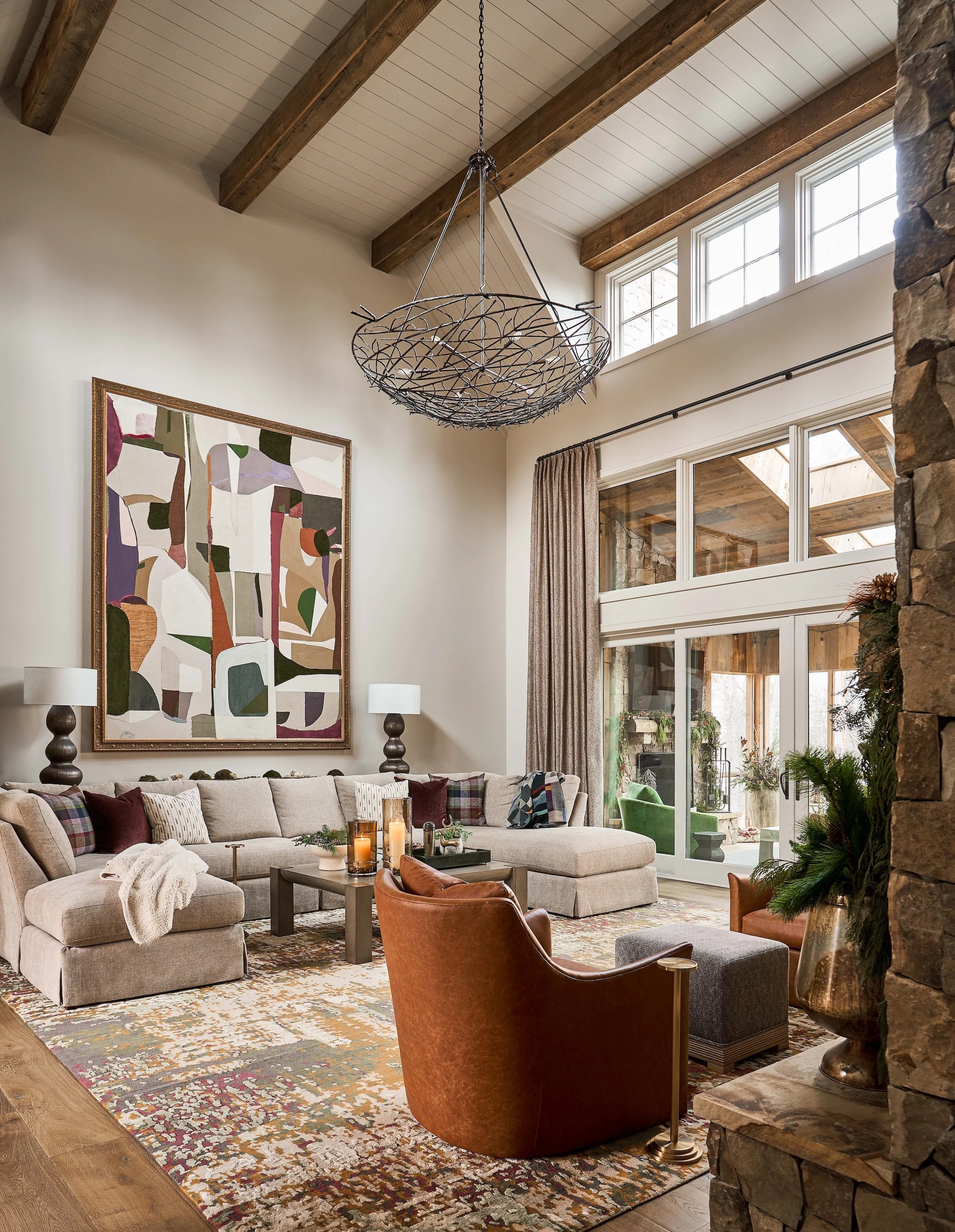 Rustic modern living room with exposed wood beams, stone fireplace, large windows, sectional sofa, leather chairs, abstract artwork, and sculptural hanging chandelier