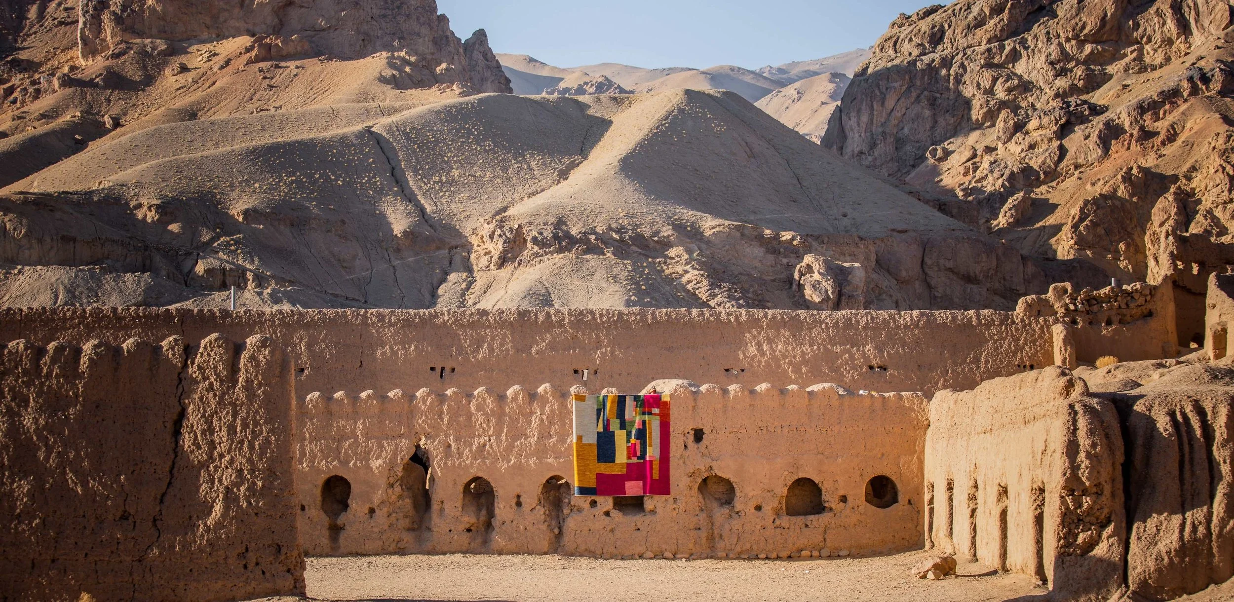 Panoramic view of rugged desert mountains behind an earthen adobe fortress wall with colorful woven textiles