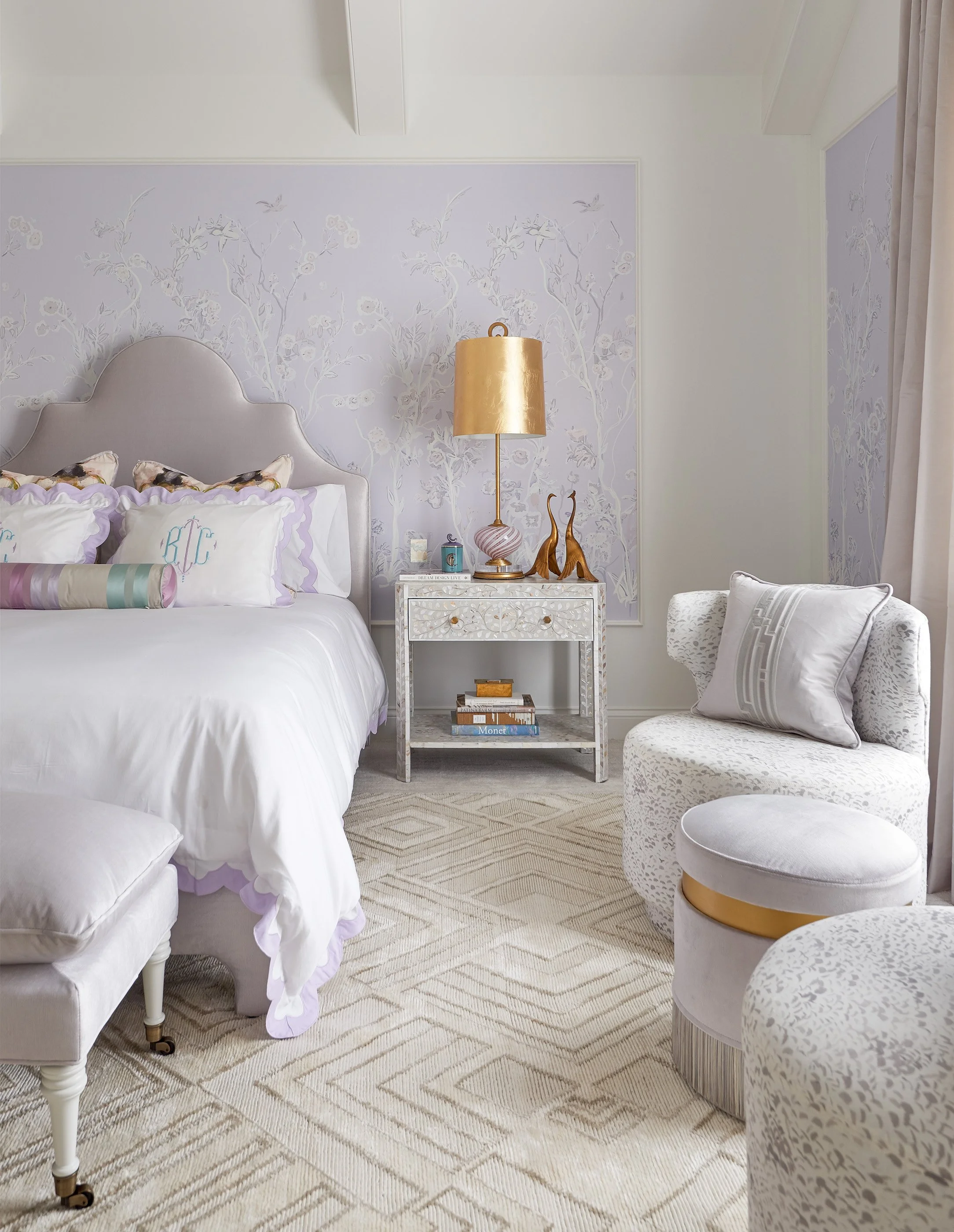Elegant pastel bedroom with floral wallpaper, upholstered bed, gold lamp, mirrored nightstand, and patterned armchair