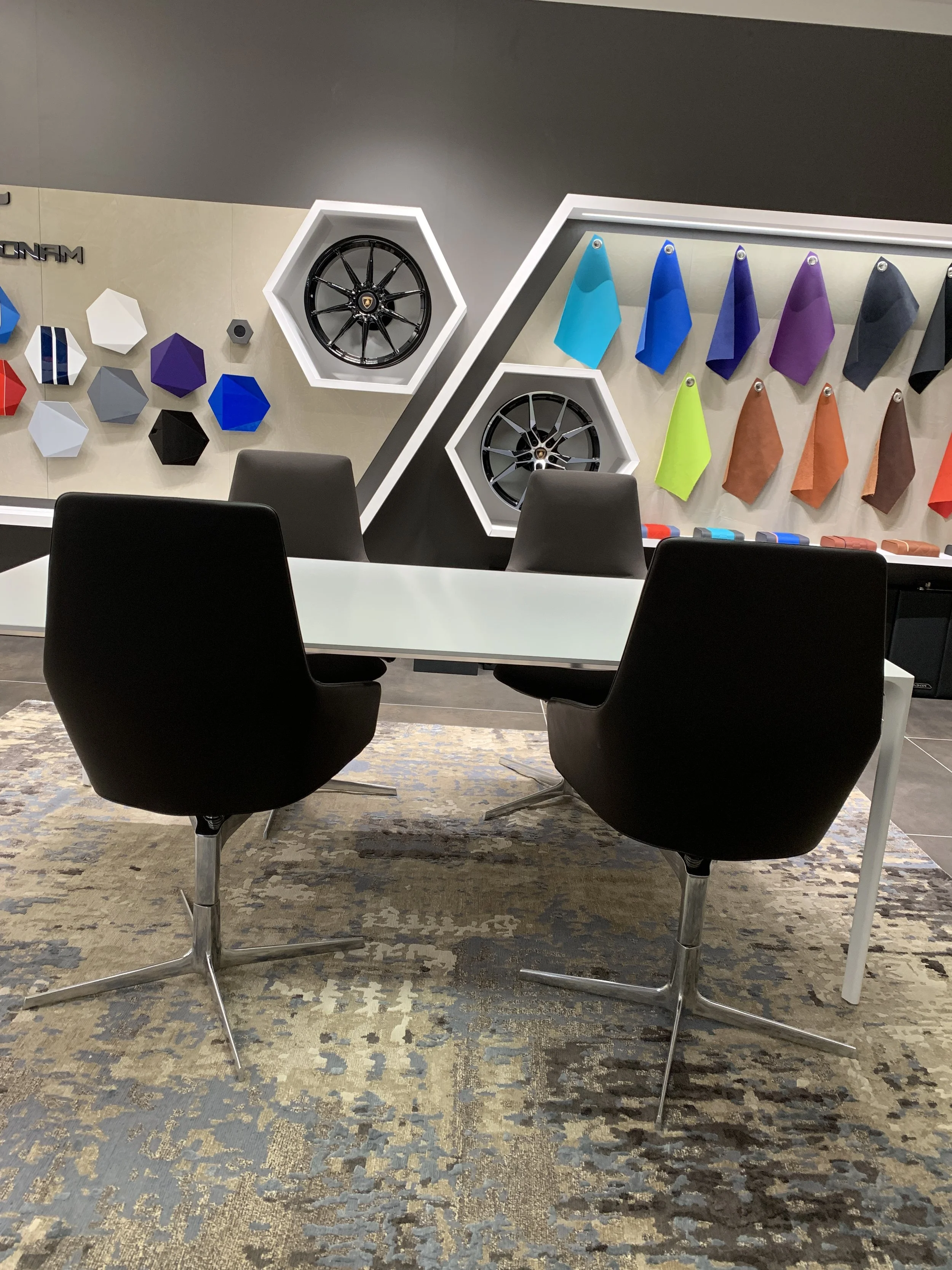 Lamborghini showroom seating area with black chairs, a white table, displayed alloy wheels, colorful leather samples, and geometric wall decor.