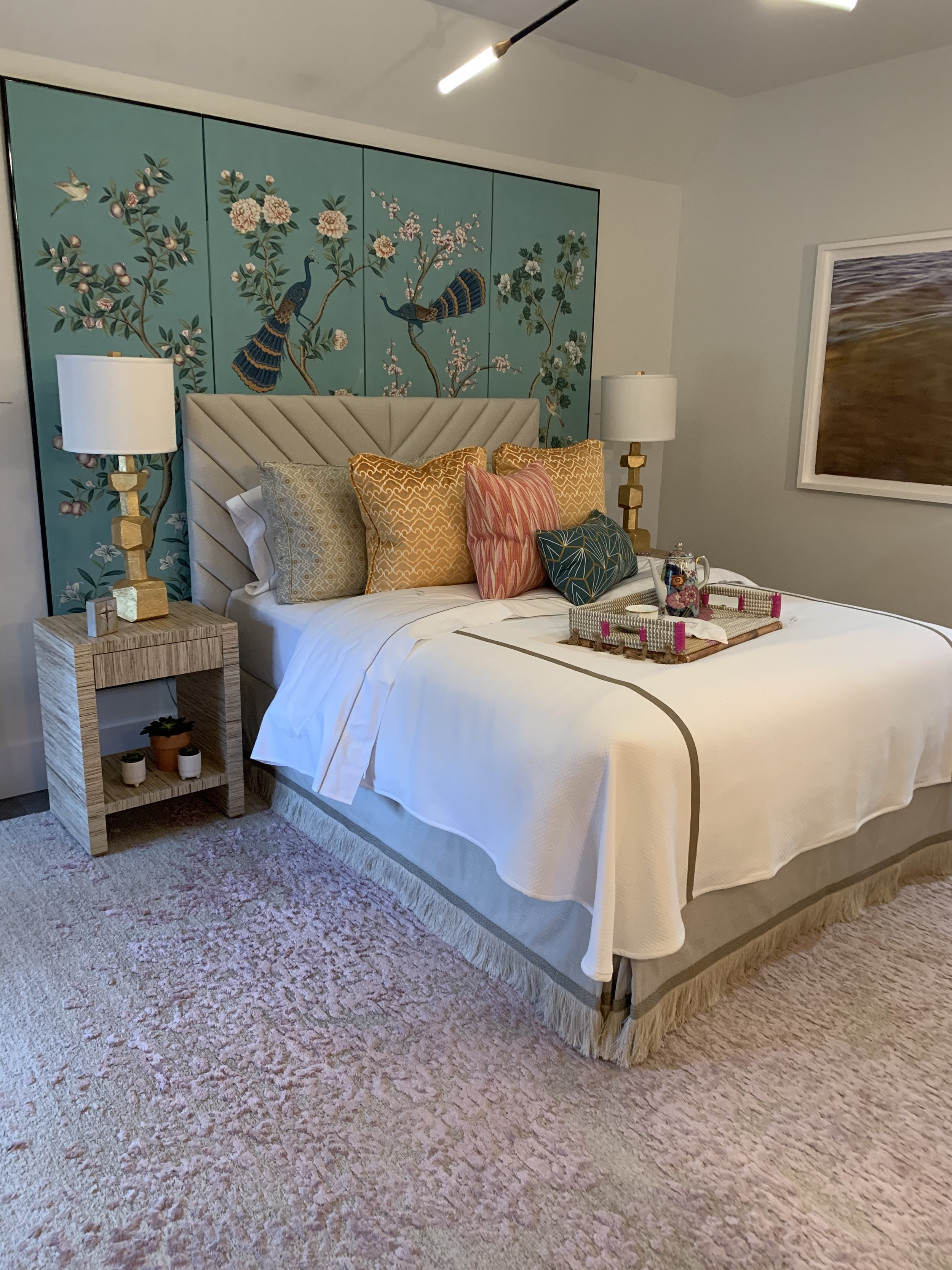 Elegant bedroom with a white bed, colorful pillows, floral peacock wall mural, gold lamps, and textured area rug