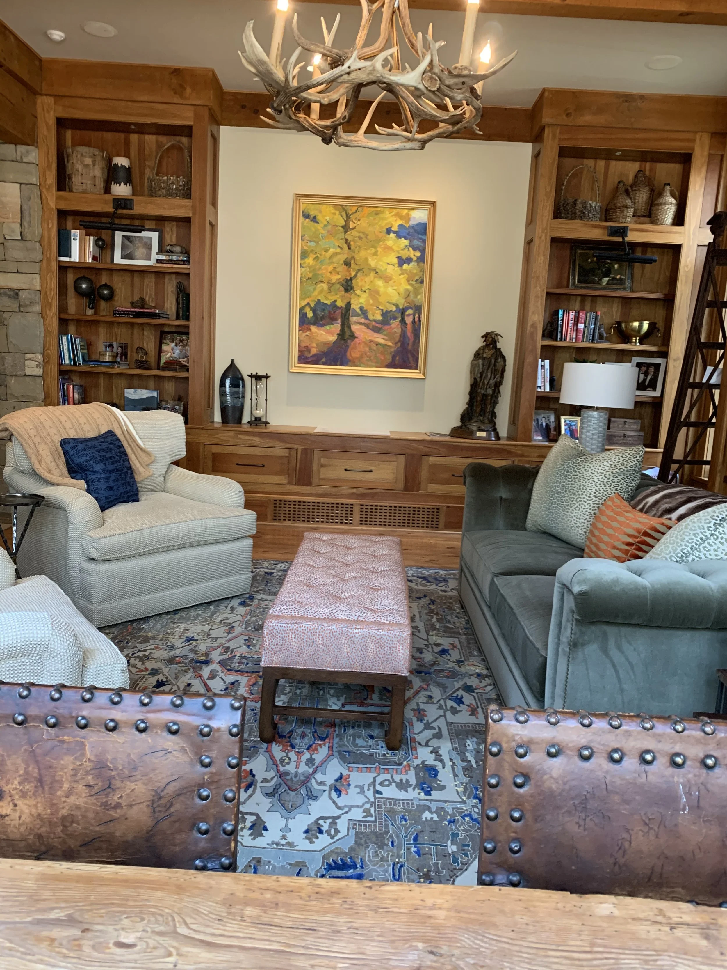 Rustic lodge living room with antler chandelier, wooden bookshelves, colorful landscape painting, upholstered seating, and patterned rug