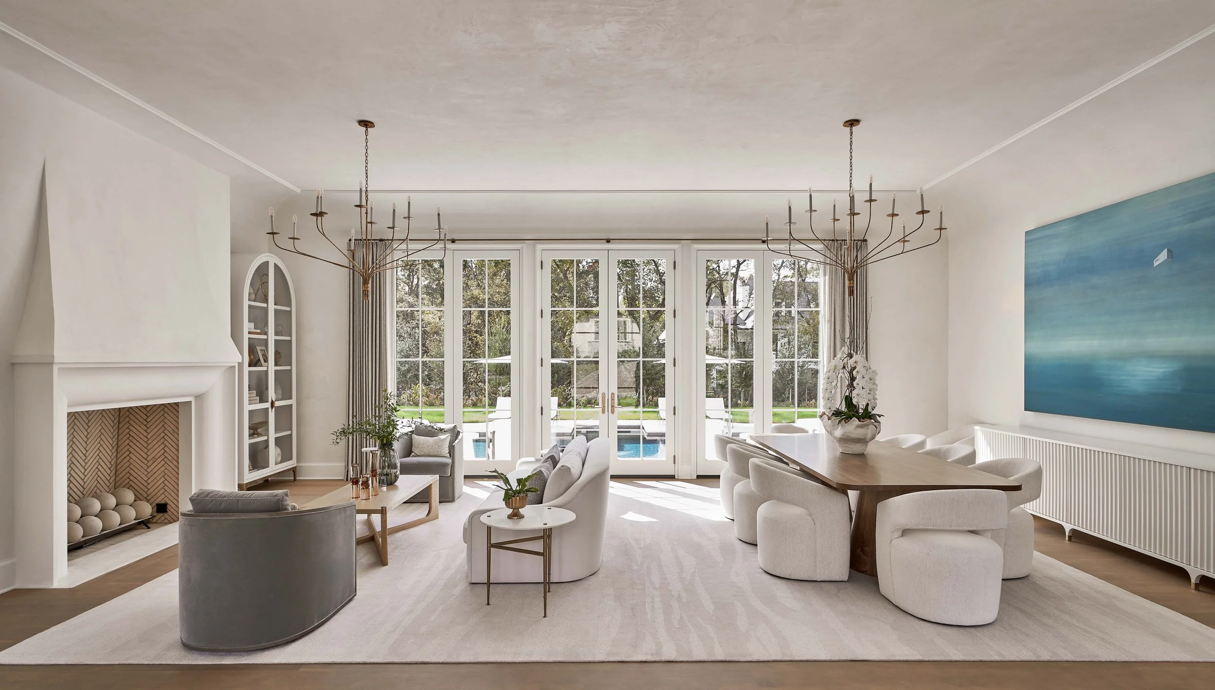 Elegant modern living and dining room with fireplace, white furnishings, chandeliers, large French doors, and backyard pool view