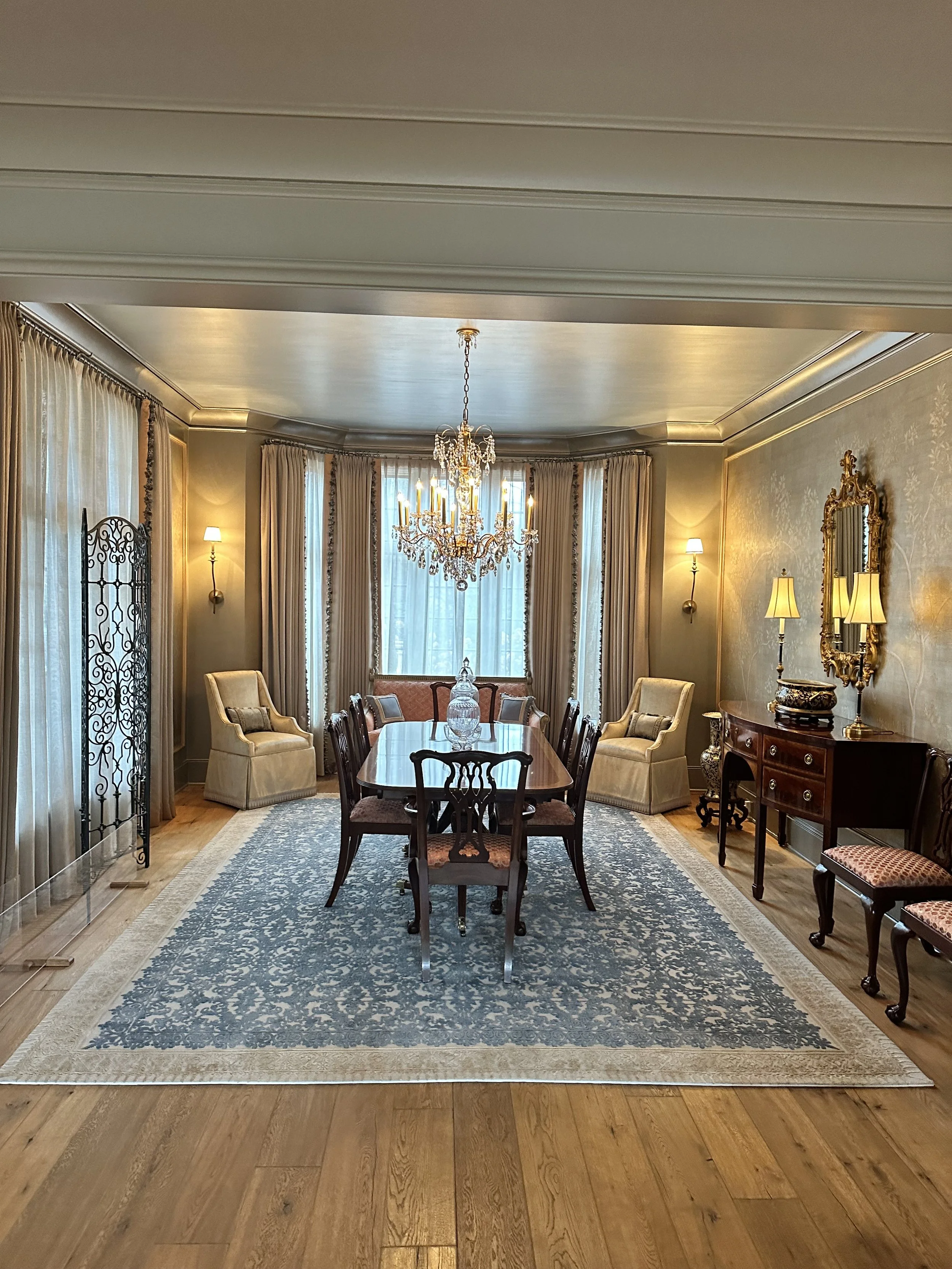 Elegant traditional dining room with crystal chandelier, long table and chairs, patterned area rug, draped windows, ornate mirror, and decorative lighting.