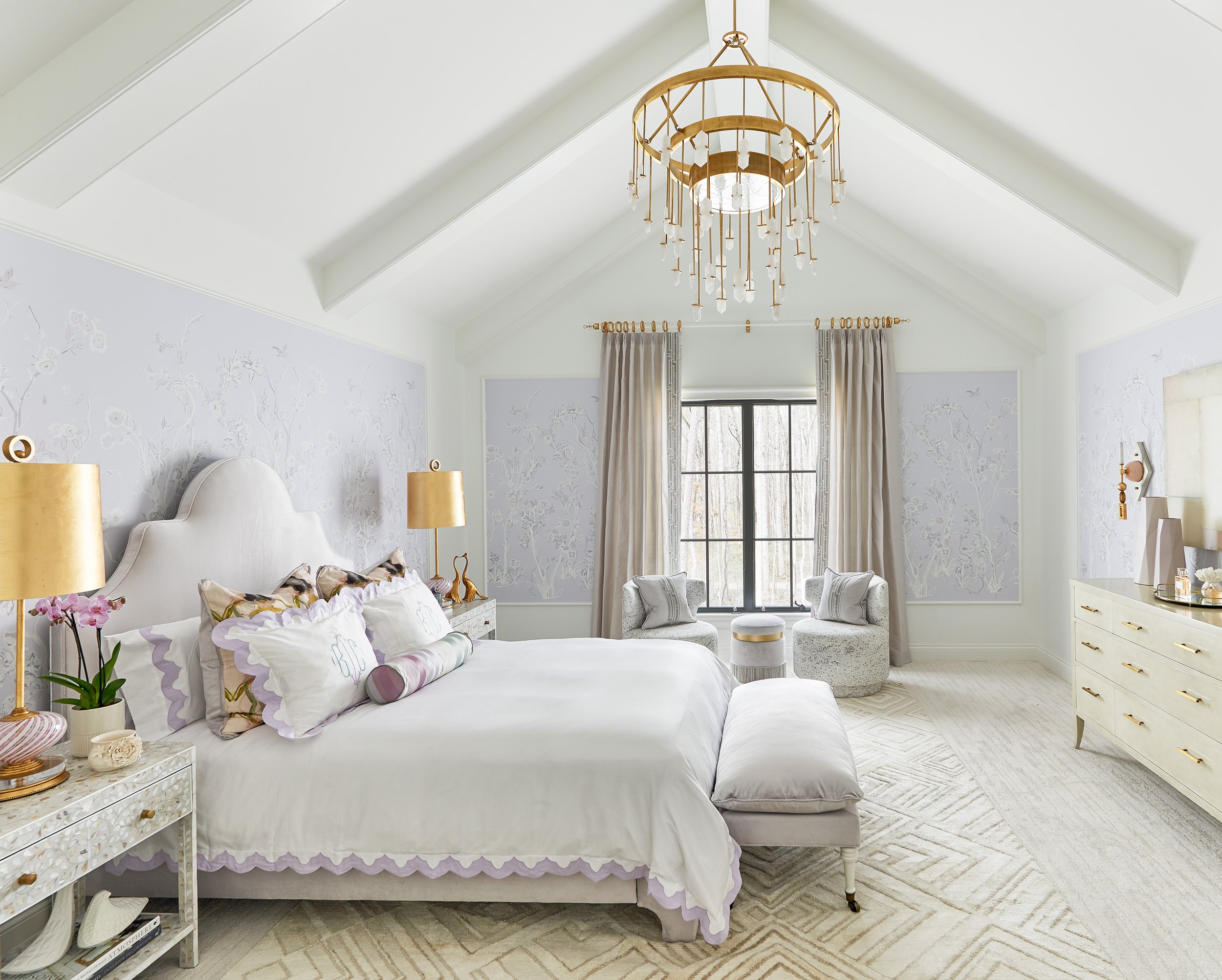 Elegant light-filled bedroom with a white upholstered bed, floral wallpaper, gold chandelier, armchairs, and large window.