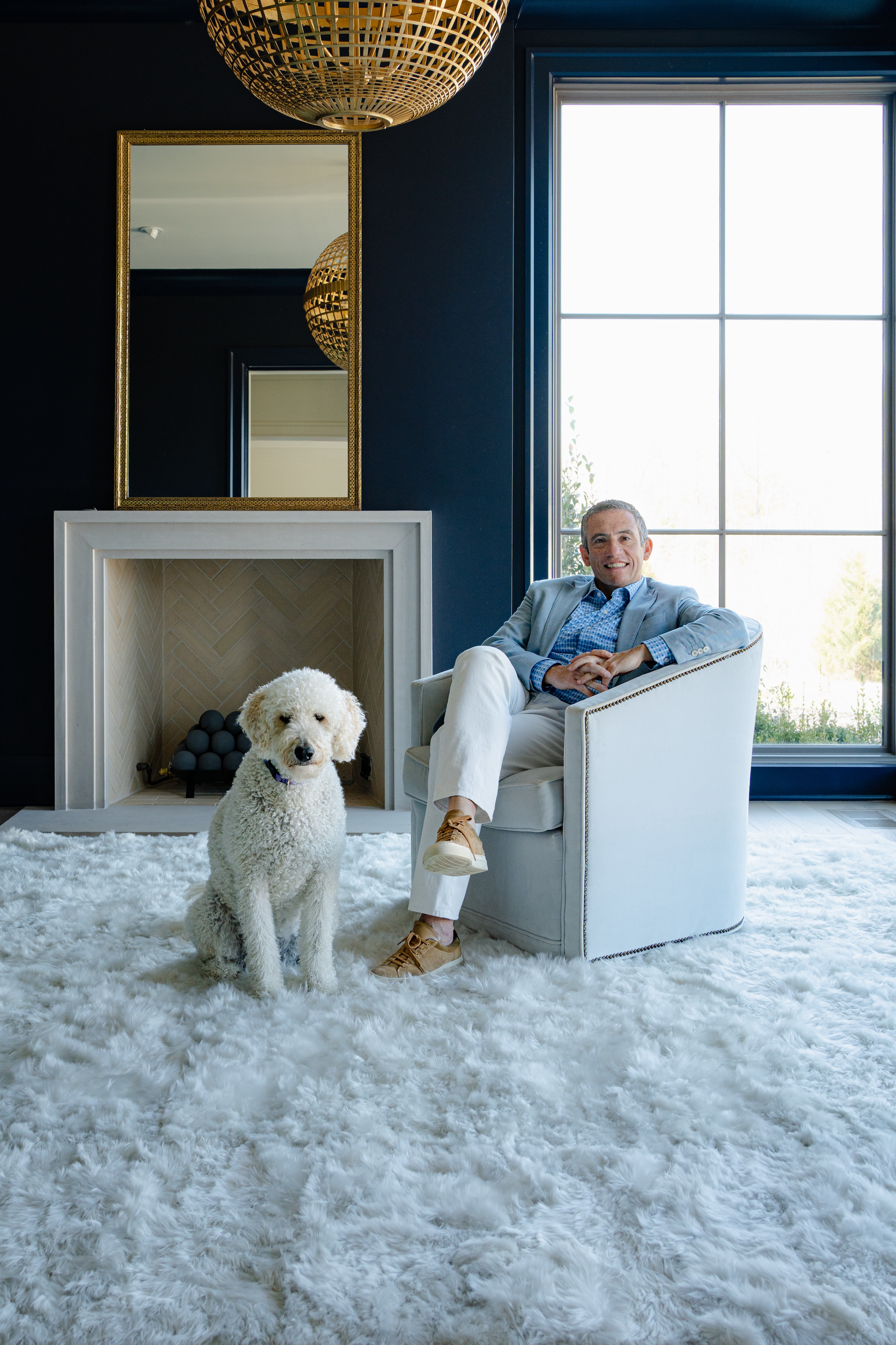 Smiling man in a light blue blazer sitting beside a white curly-haired dog in a stylish living room.