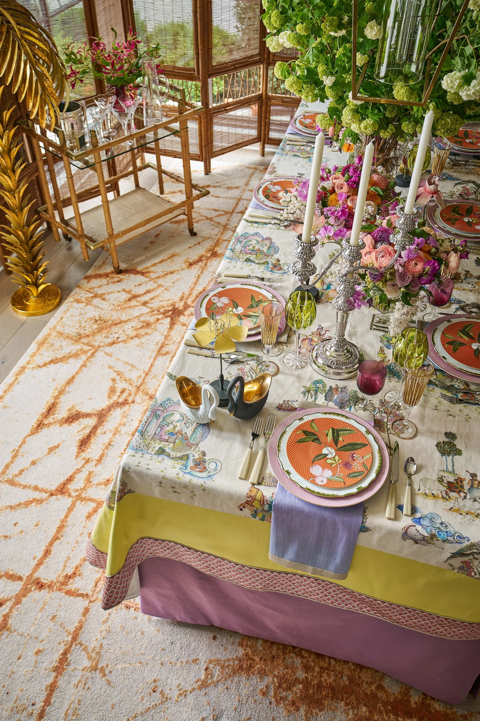 Colorful floral dining table setting with patterned plates, silver candlesticks, flowers, glassware, and bamboo chairs in a sunroom