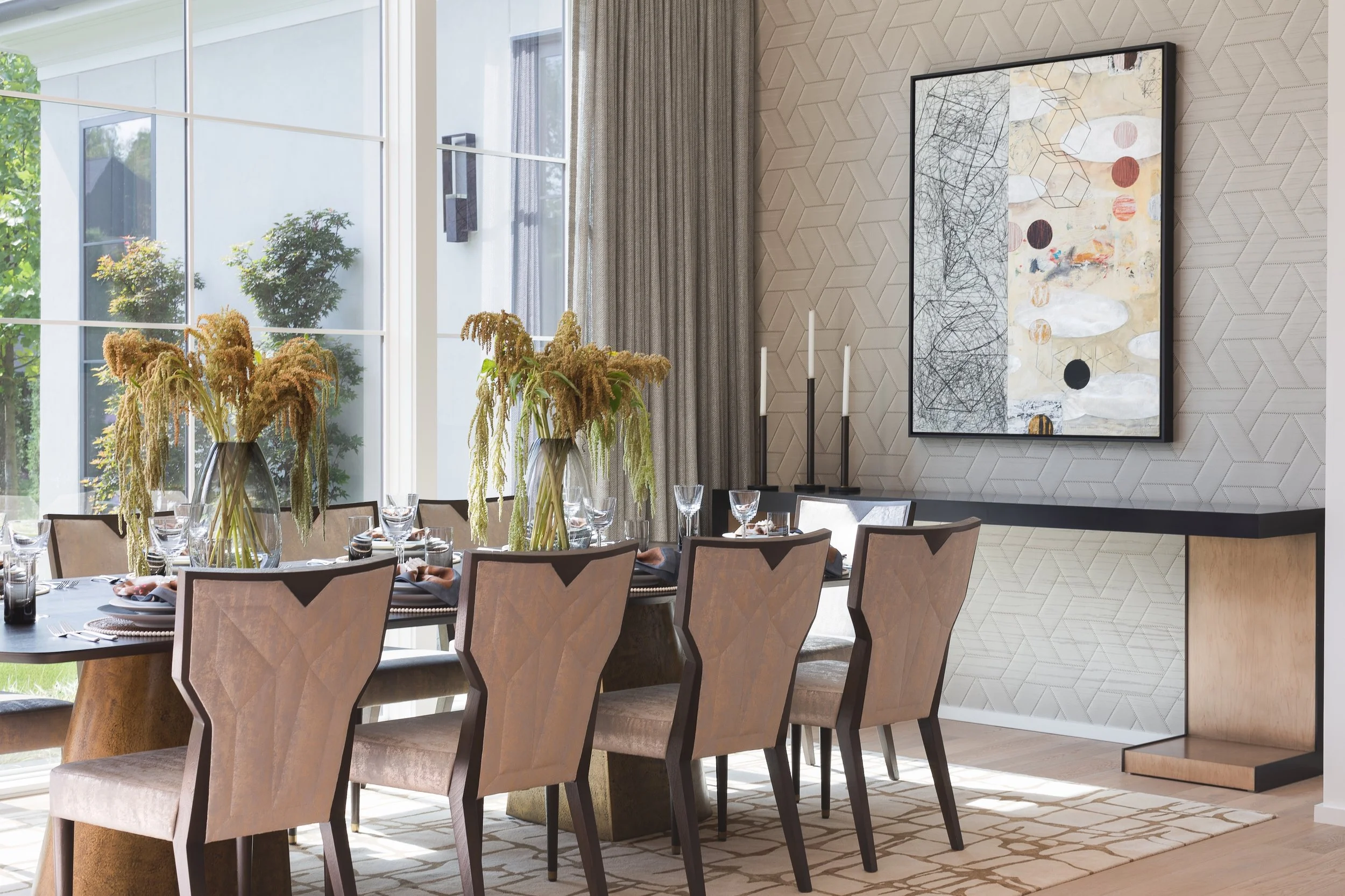Elegant dining room with a set table, upholstered chairs, floral centerpieces, large windows, geometric wallpaper, and abstract wall art