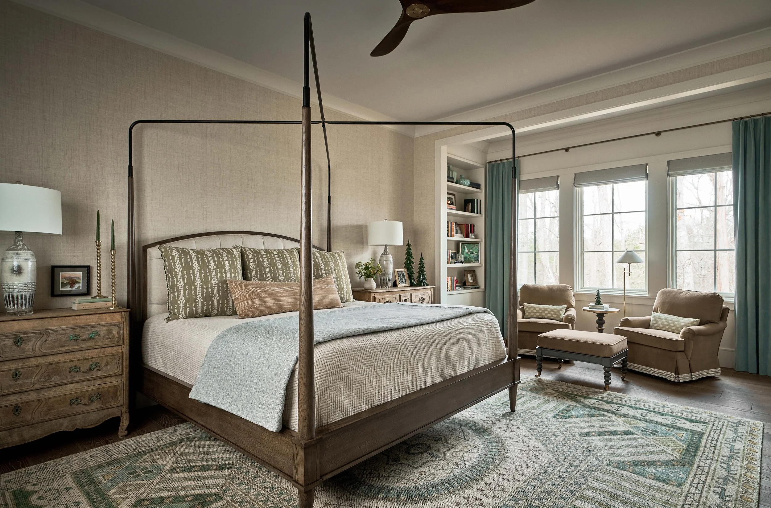 Elegant bedroom with a dark wood four-poster bed, patterned bedding, dresser, reading chairs, built-in bookshelves, and large windows with teal curtains