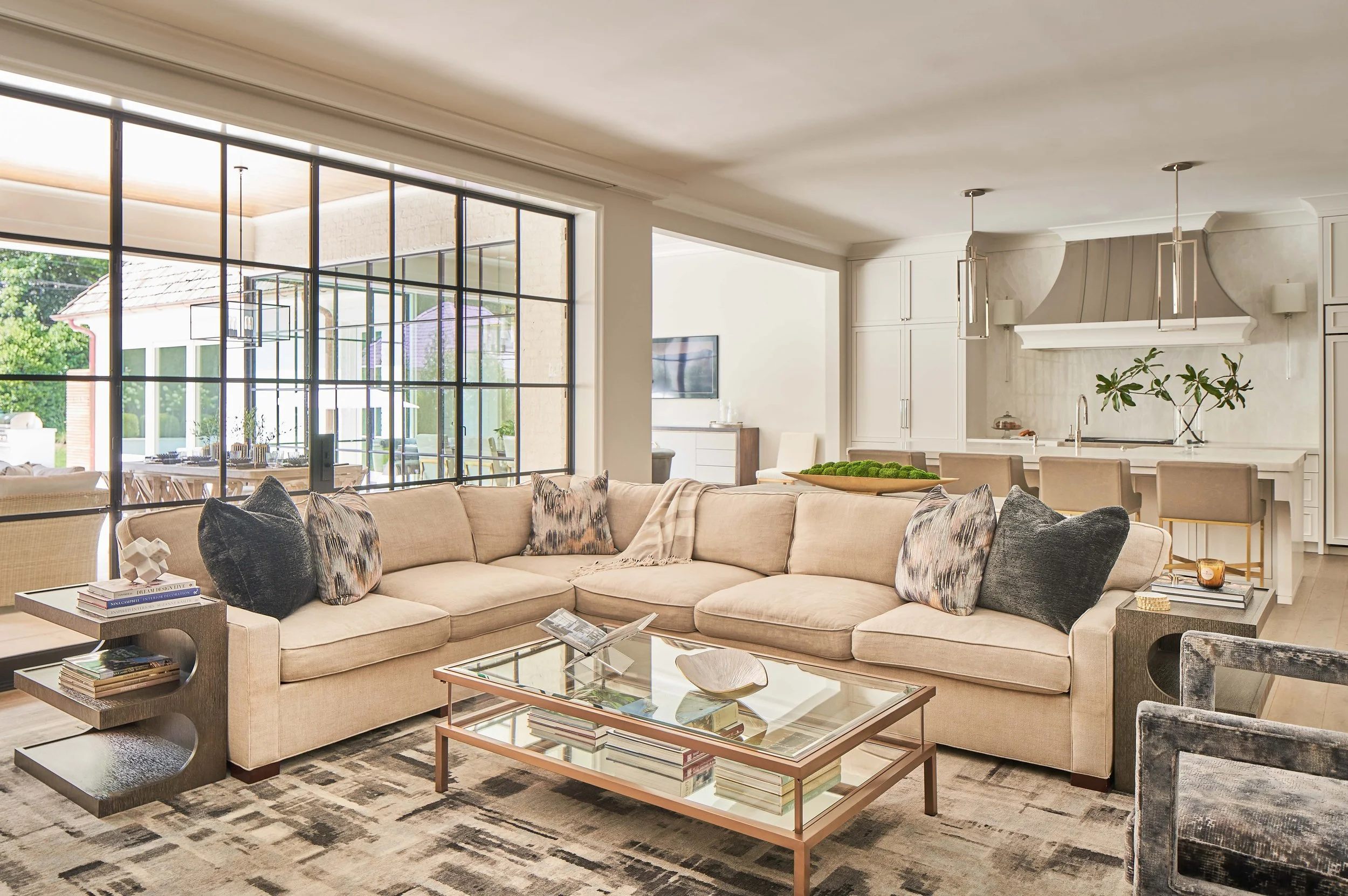 Elegant open-concept living room and kitchen with a sectional sofa, glass coffee table, large black-framed windows, and island seating