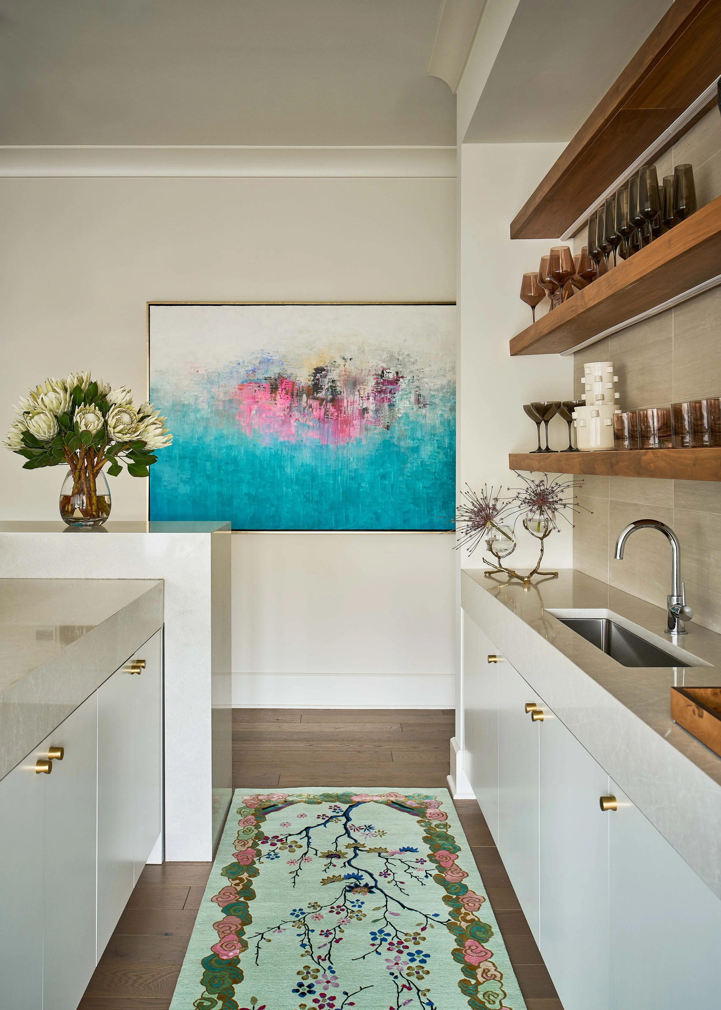 Modern white kitchen with wood shelves, glassware, floral arrangement, abstract turquoise artwork, and patterned runner rug