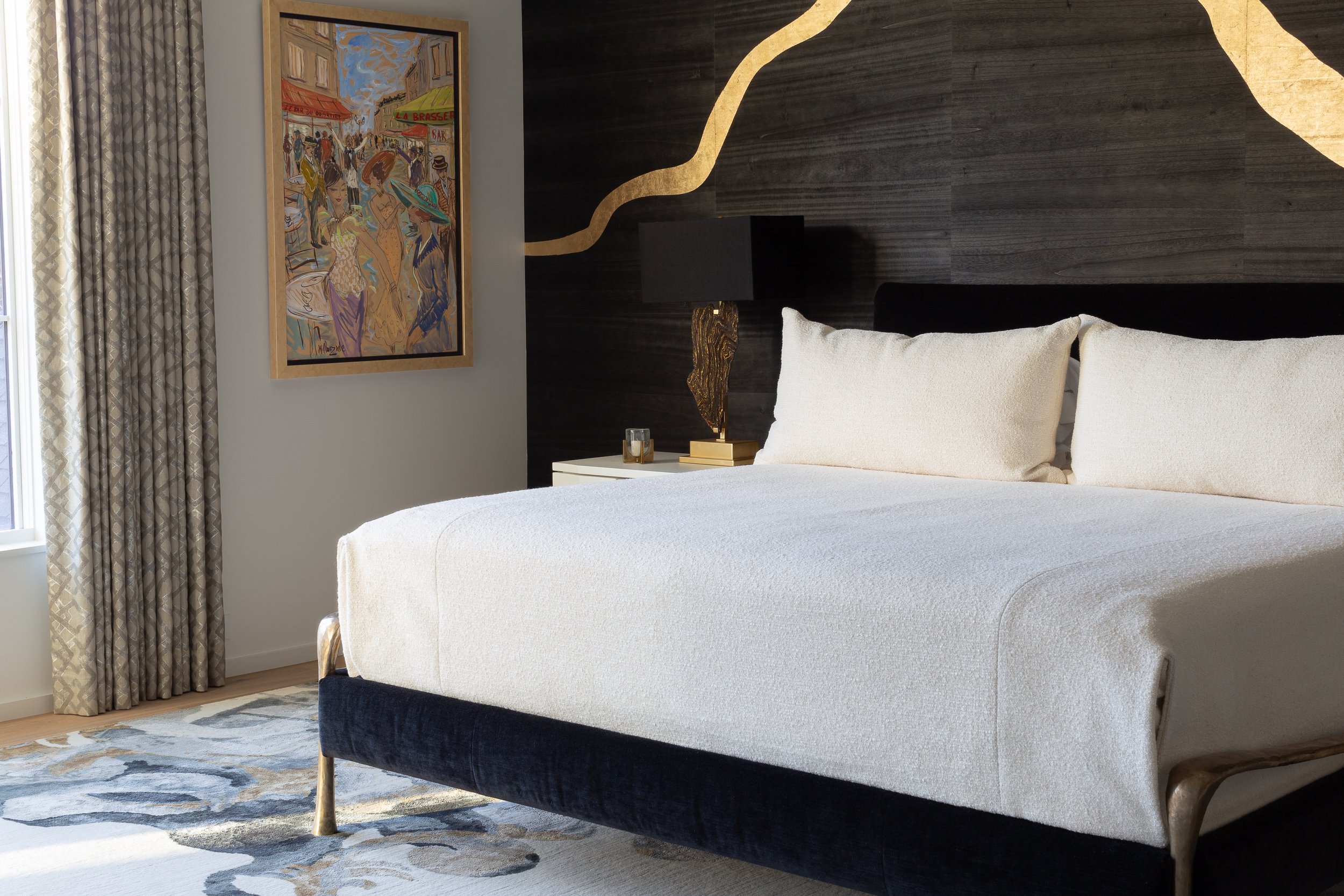 Elegant bedroom with a white upholstered bed, navy velvet frame, black wood accent wall, bedside lamp, framed artwork, and patterned curtains.