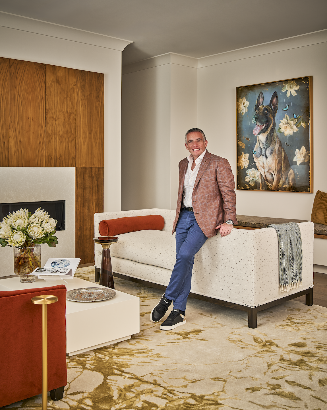 Smiling man in a plaid blazer standing in a stylish living room beside a cream sofa and dog portrait
