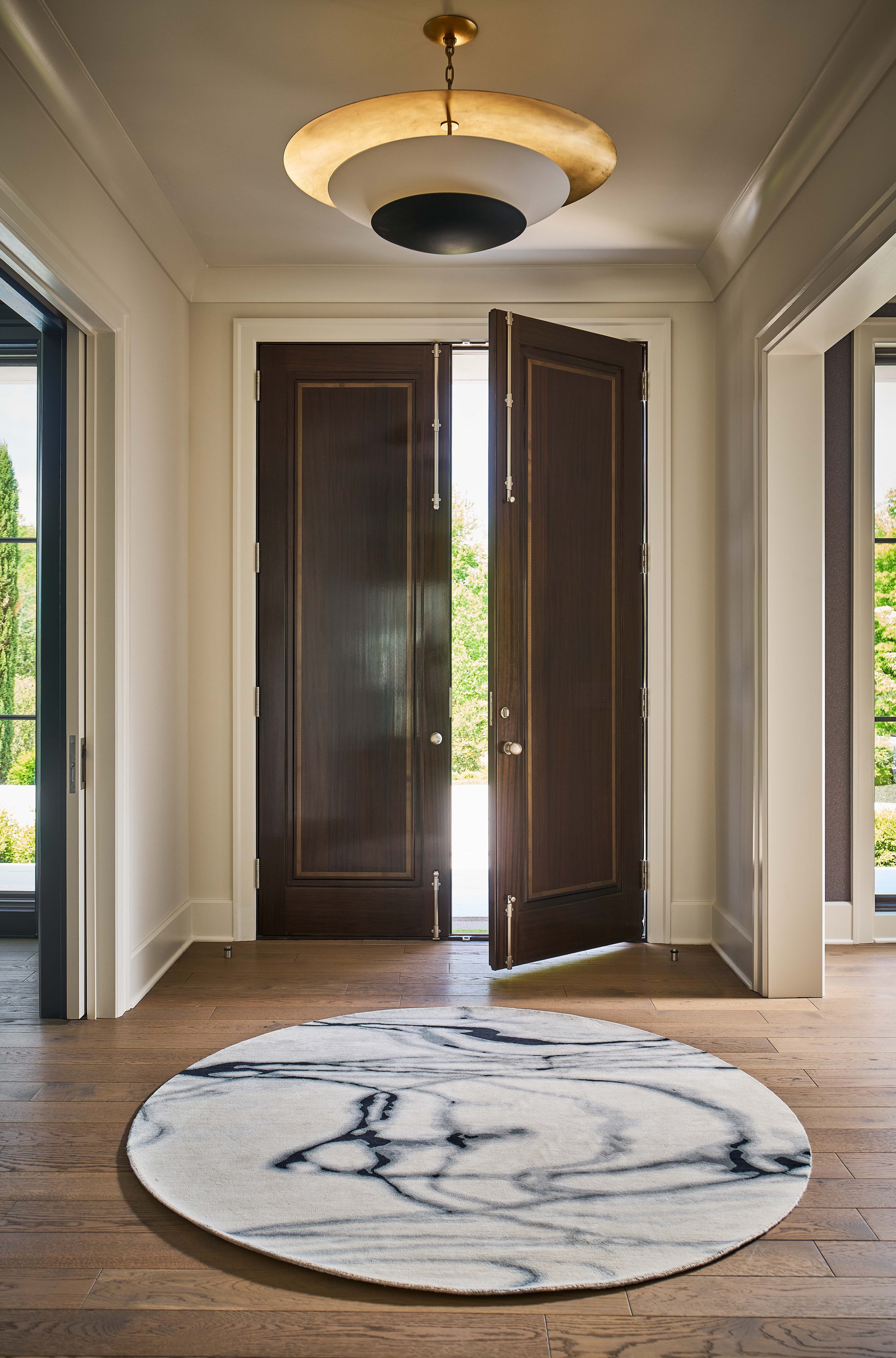 Elegant entryway with open dark wood double doors, a gold pendant light, hardwood floors, and a round marble-patterned rug