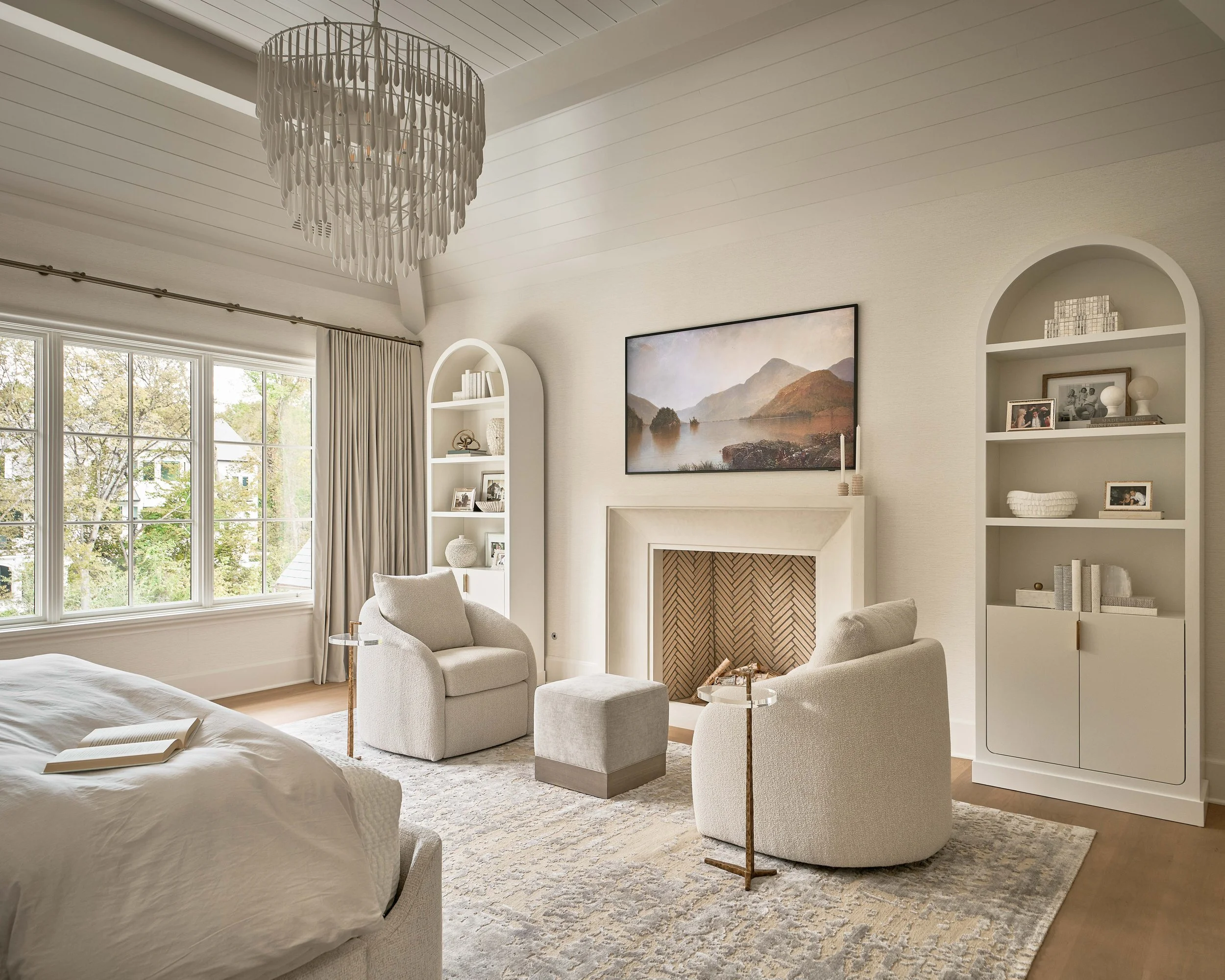 Elegant neutral-toned bedroom sitting area with a fireplace, mountain landscape artwork, arched built-in shelves, chandelier, bed, and two upholstered chairs
