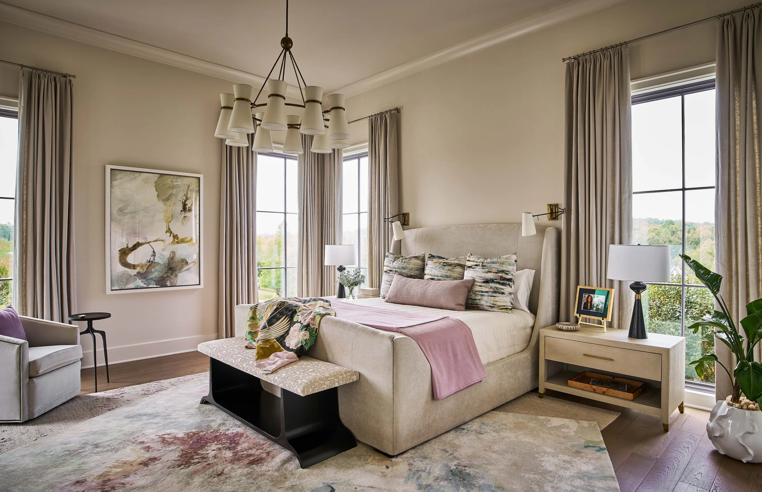 Elegant bedroom with upholstered bed, patterned bench, chandelier, large windows, neutral curtains, bedside tables, and indoor plants