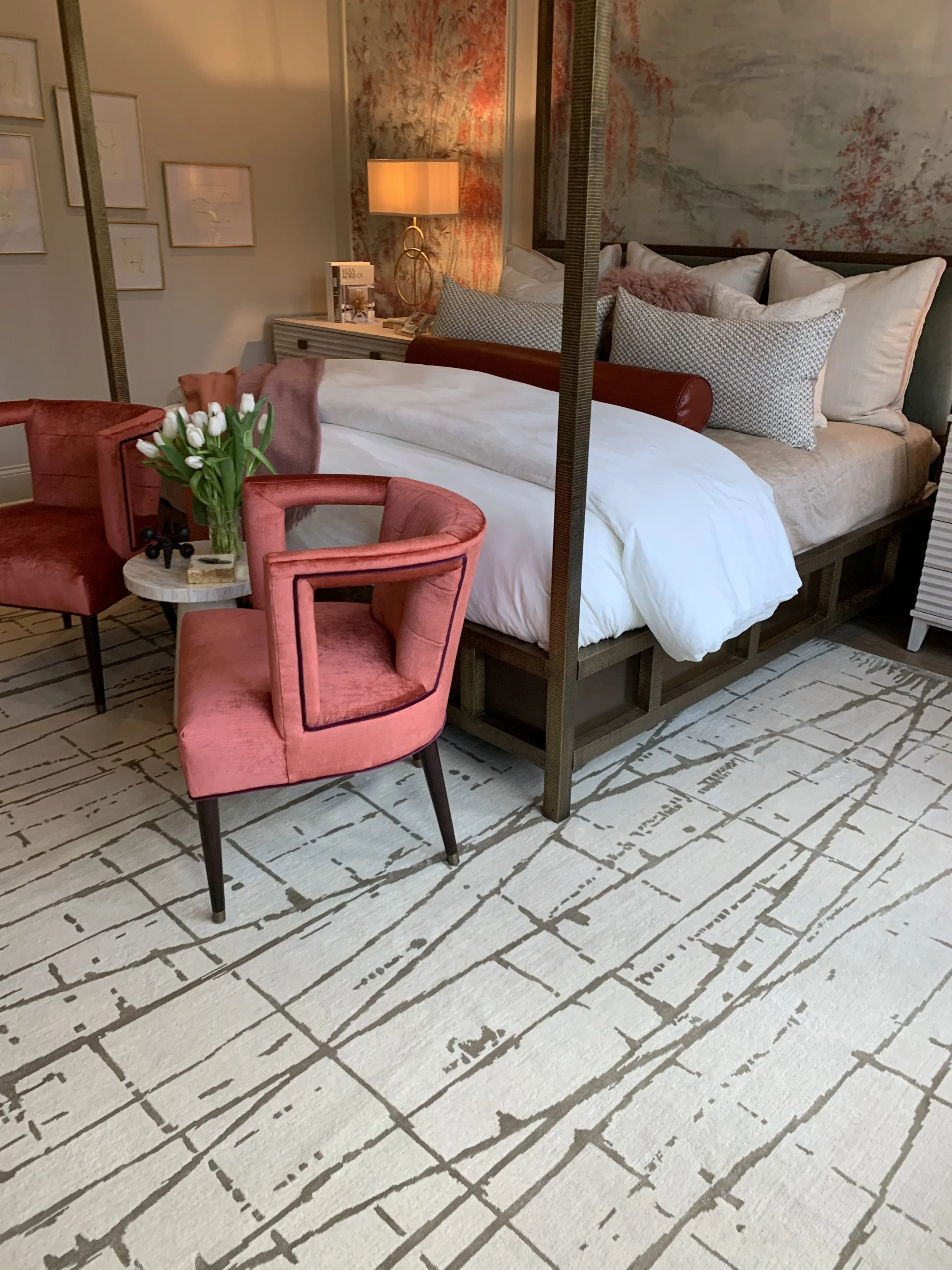 Elegant bedroom with a four-poster bed, white bedding, patterned pillows, coral armchairs, bedside lamp, floral wallpaper, and geometric area rug