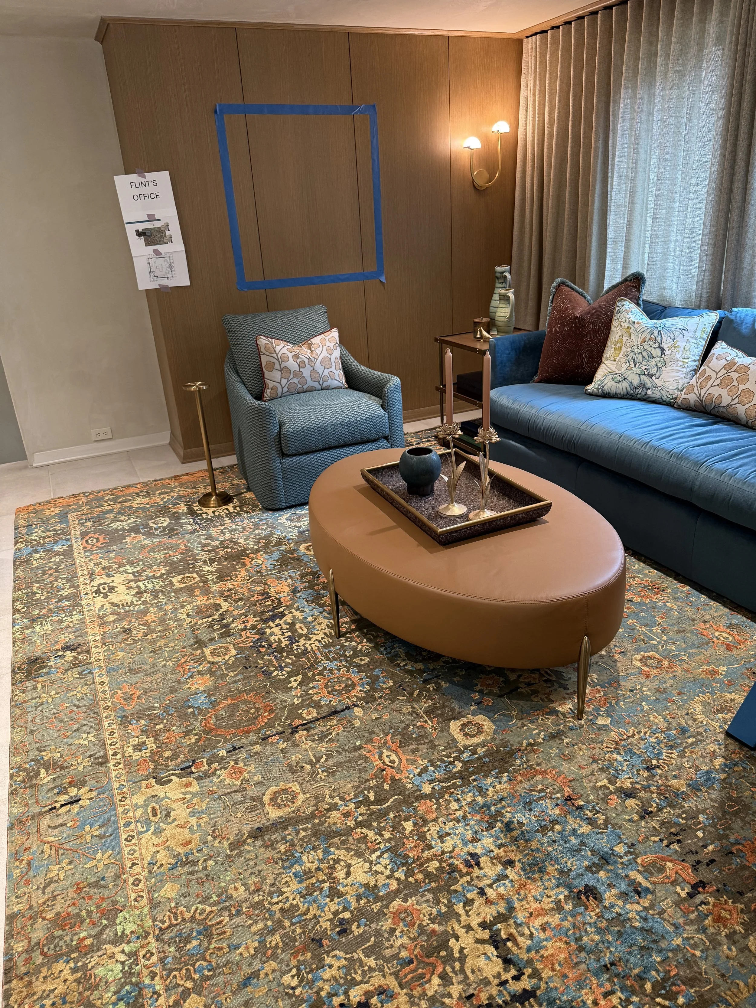 Colorful modern living room with blue sofa, patterned armchair, round leather ottoman, wood-paneled wall, floor lamp, curtains, and patterned area rug.