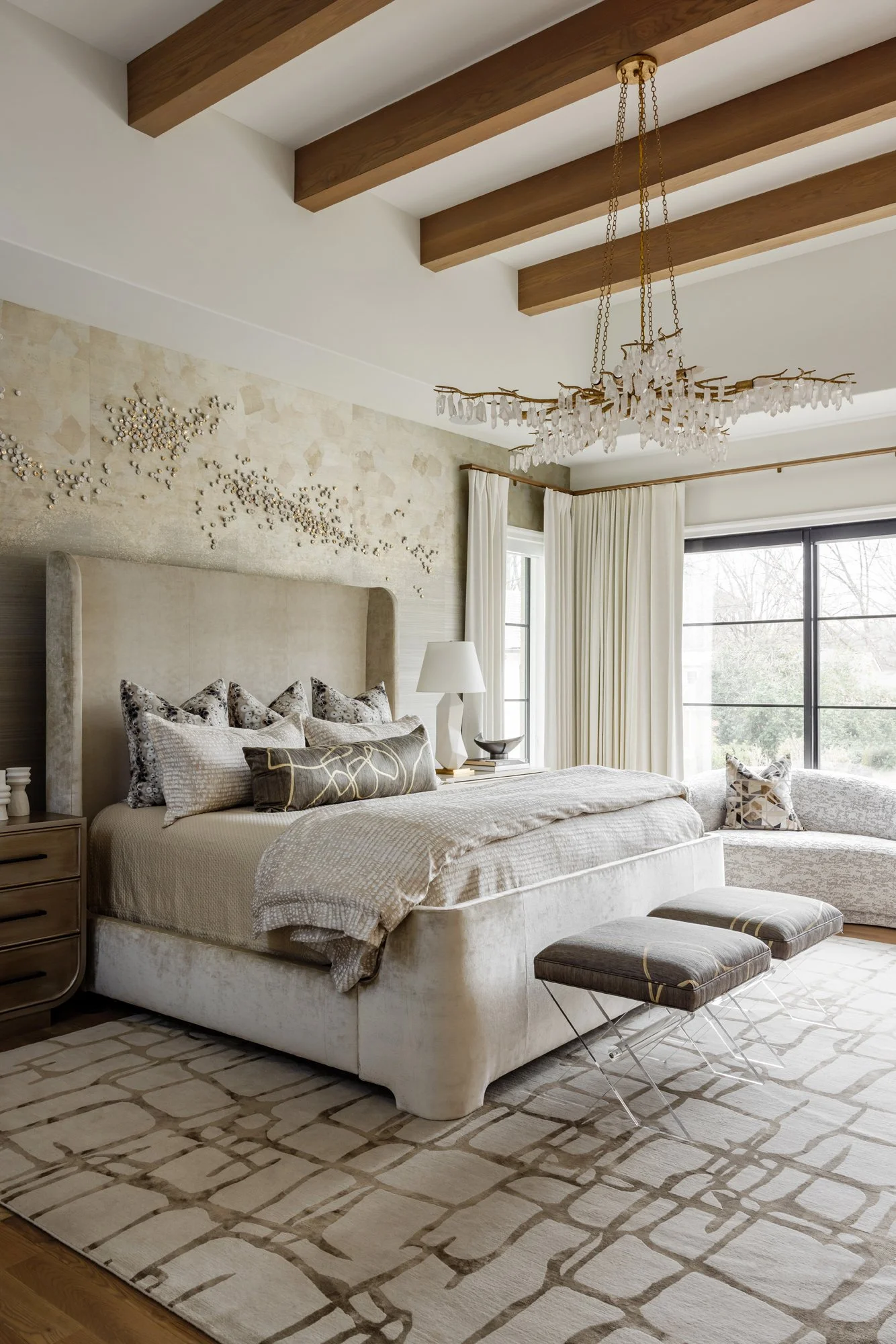 Elegant neutral-toned bedroom with a plush upholstered bed, crystal chandelier, exposed wood ceiling beams, patterned rug, and large windows.