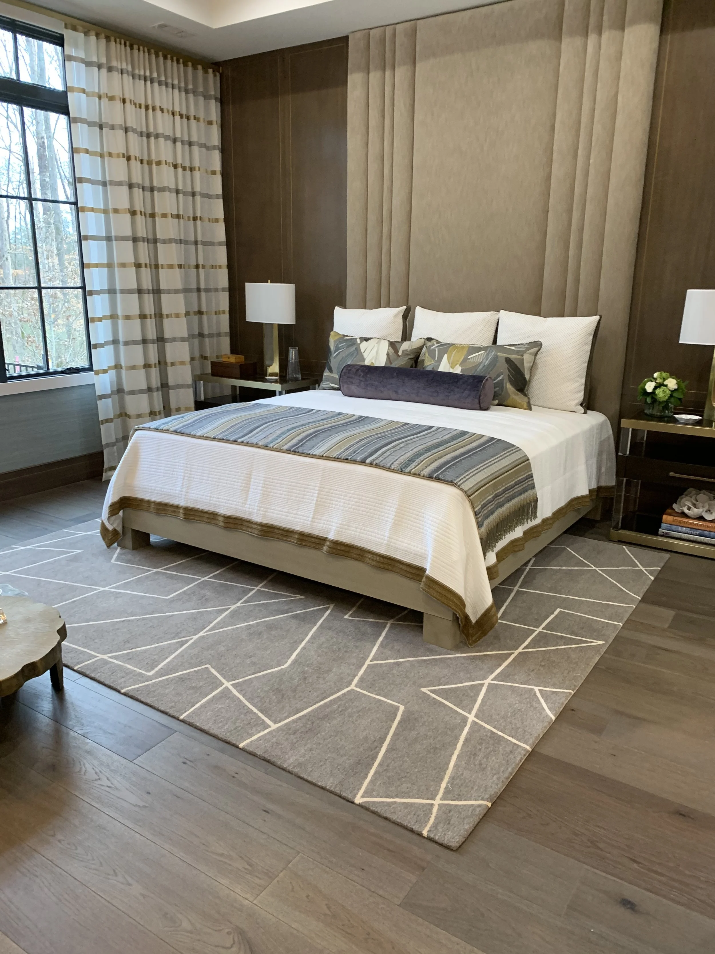 Modern bedroom with a neatly made bed, patterned throw, decorative pillows, bedside lamps, wood-paneled accent wall, and large window.