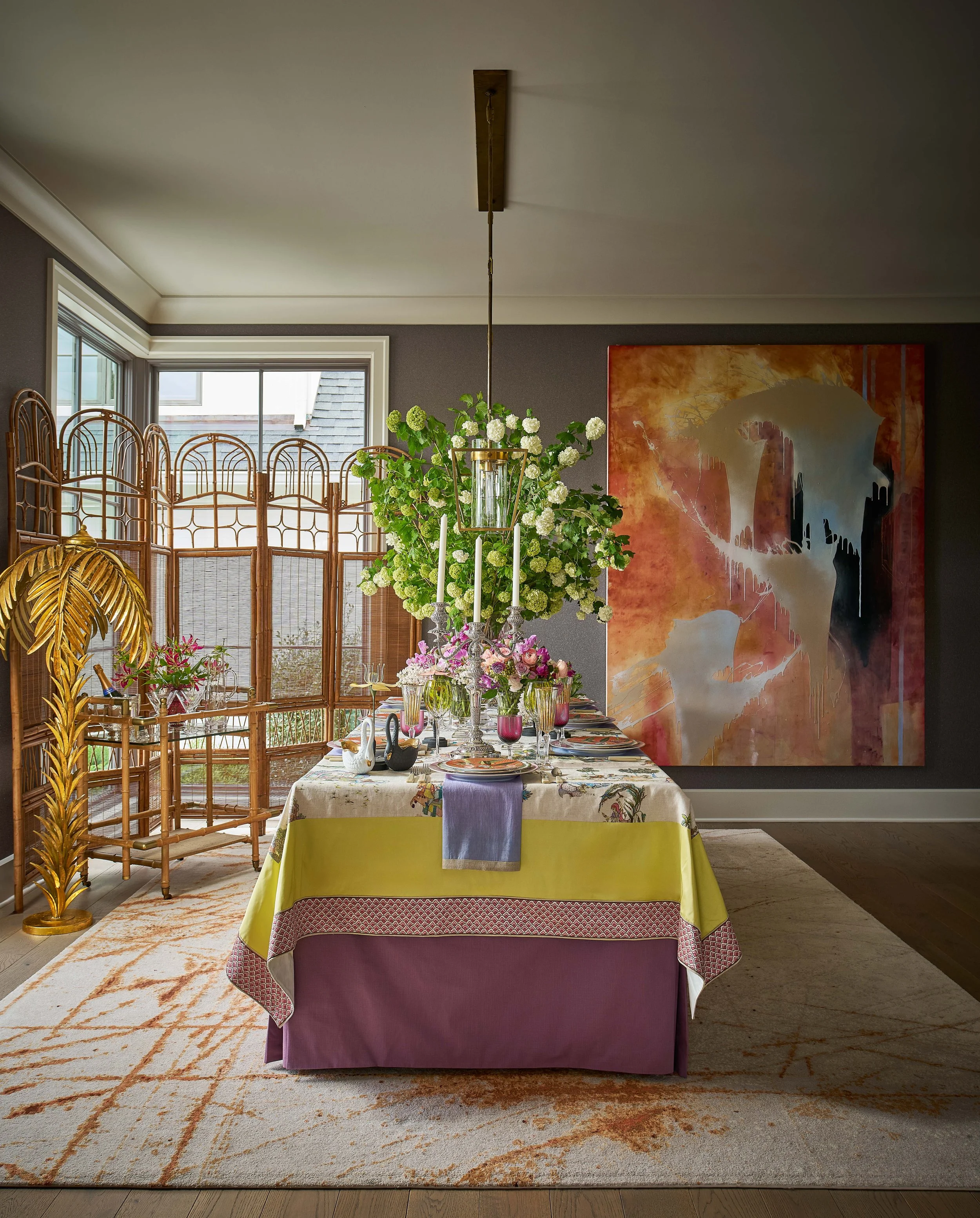 Colorful dining room with a set table, floral centerpiece, wicker chairs, large abstract artwork, and pendant light
