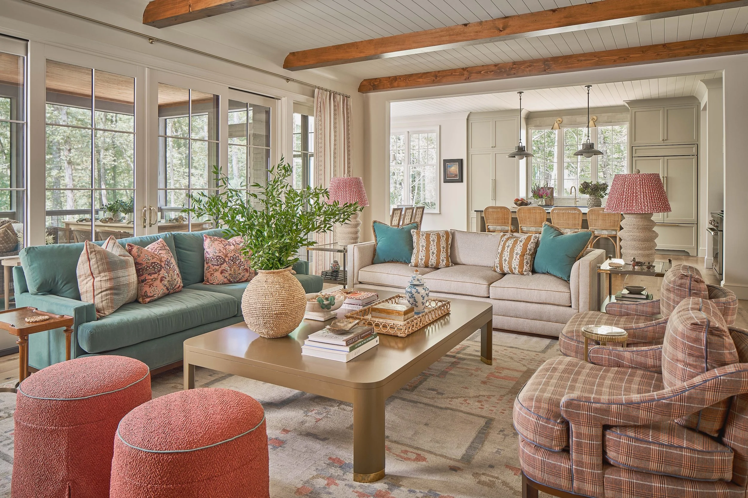 Cozy open-concept living room and kitchen with exposed wood beams, teal and beige sofas, plaid armchairs, coffee table, plants, and large windows overlooking trees