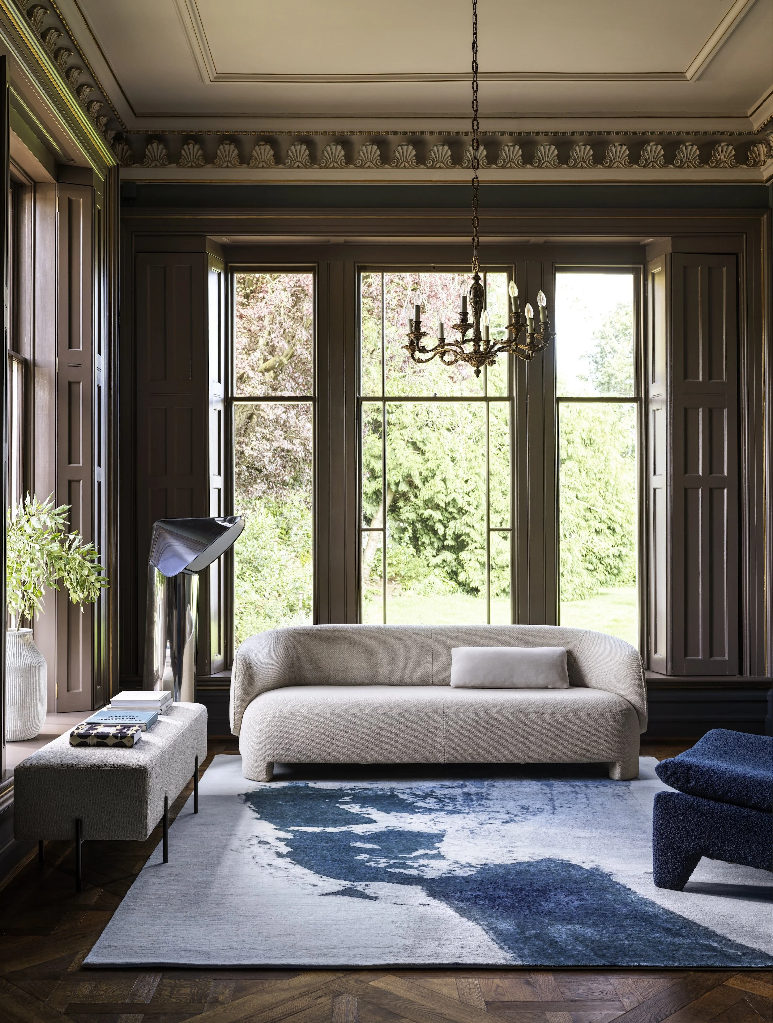 Elegant living room with a cream sofa, ornate chandelier, dark wood paneling, large garden-view windows, and a blue abstract area rug.