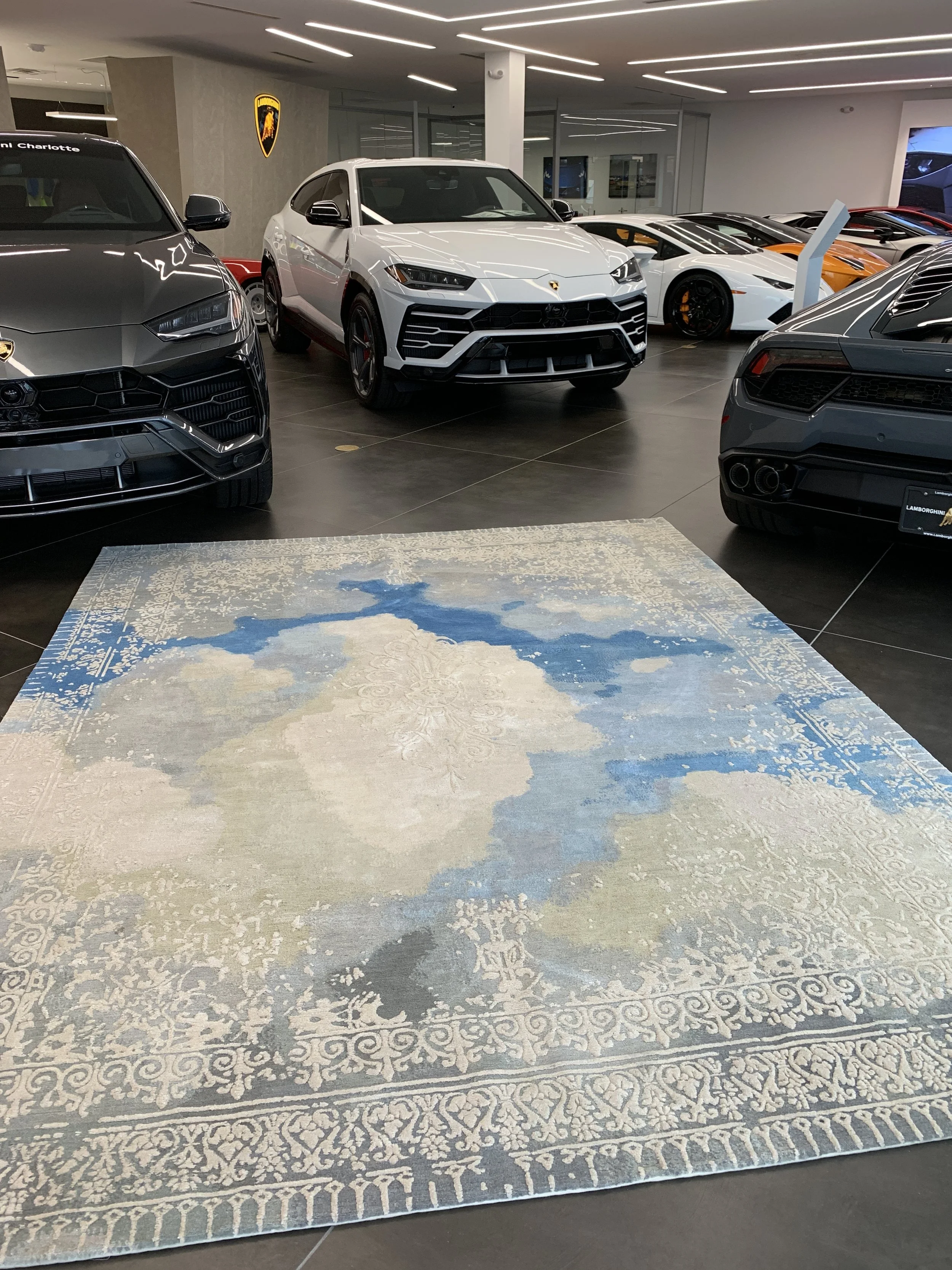 Luxury Lamborghini showroom displaying a white Urus SUV and several colorful sports cars on a patterned rug.