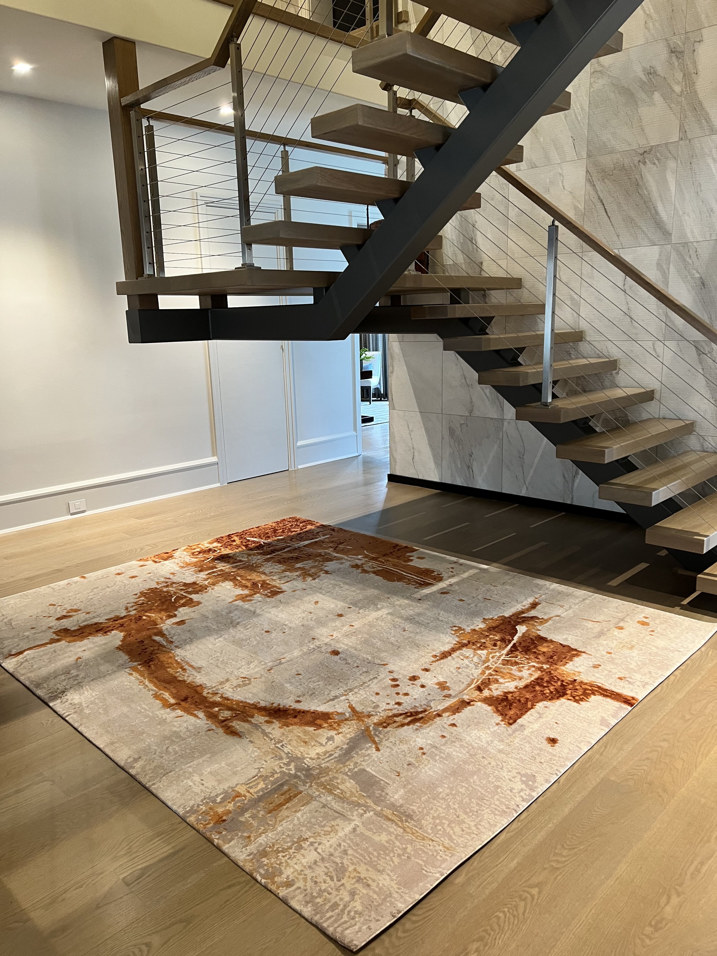 Modern floating wooden staircase with black steel supports and cable railing beside a marble-clad wall and patterned area rug