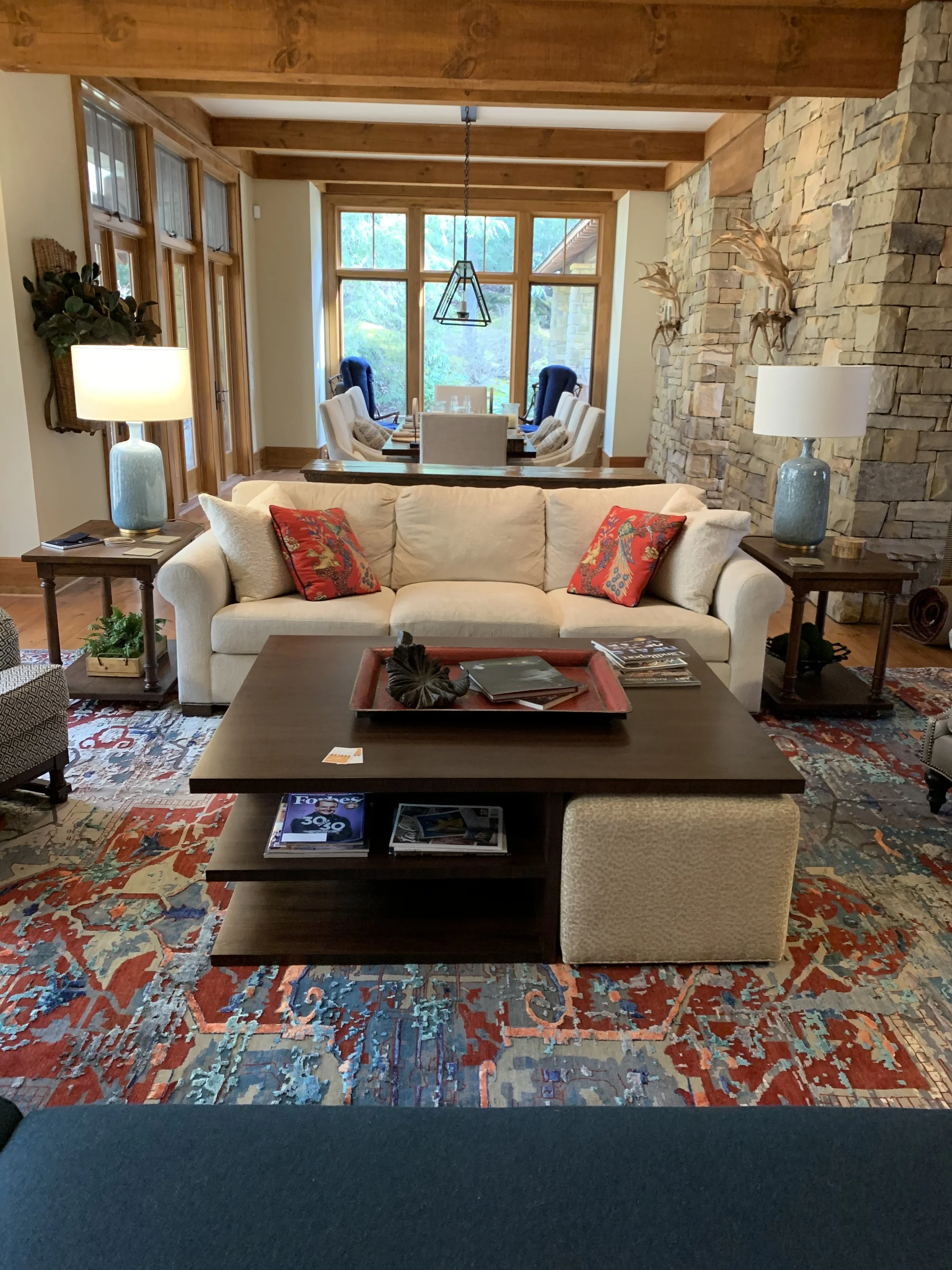 Rustic living room with exposed wood beams, stone fireplace, cream sofa, colorful pillows, coffee table, and patterned rug