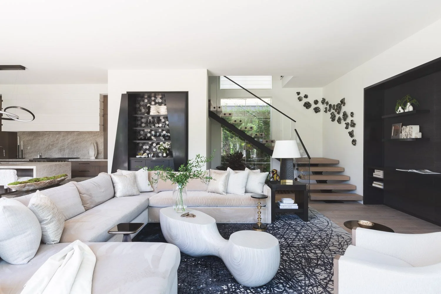 Modern open-concept living room with white sectional sofa, sculptural coffee table, black patterned rug, contemporary staircase, and adjacent kitchen