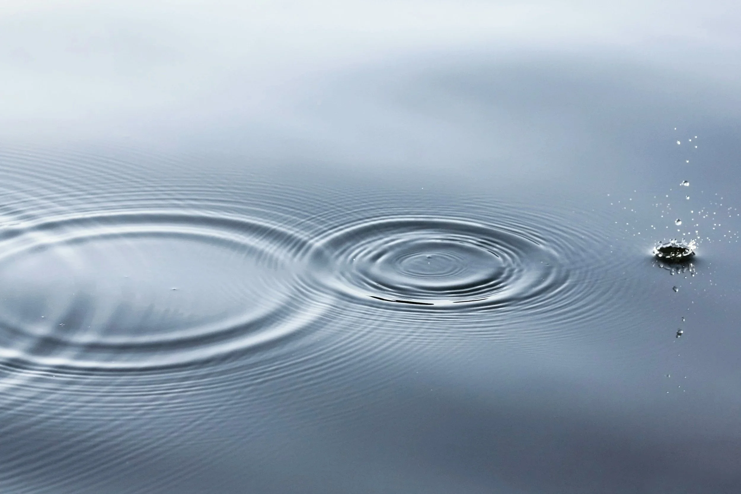 Close-up of water with ripples and droplet splash, reflecting a cloudy sky.