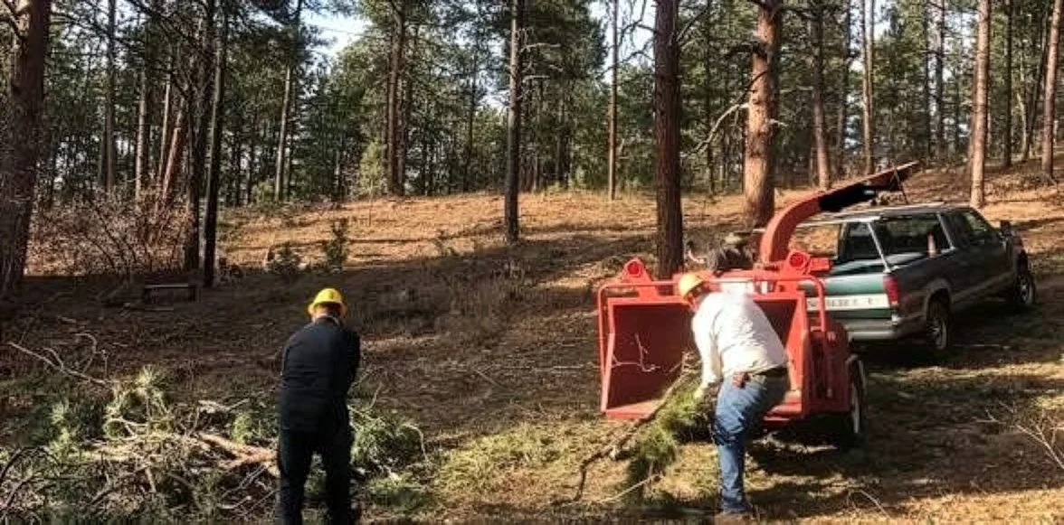 Two workers in yellow helmets and dark clothing are operating a wood chipper in a forested area, with a car with an open trunk parked nearby.