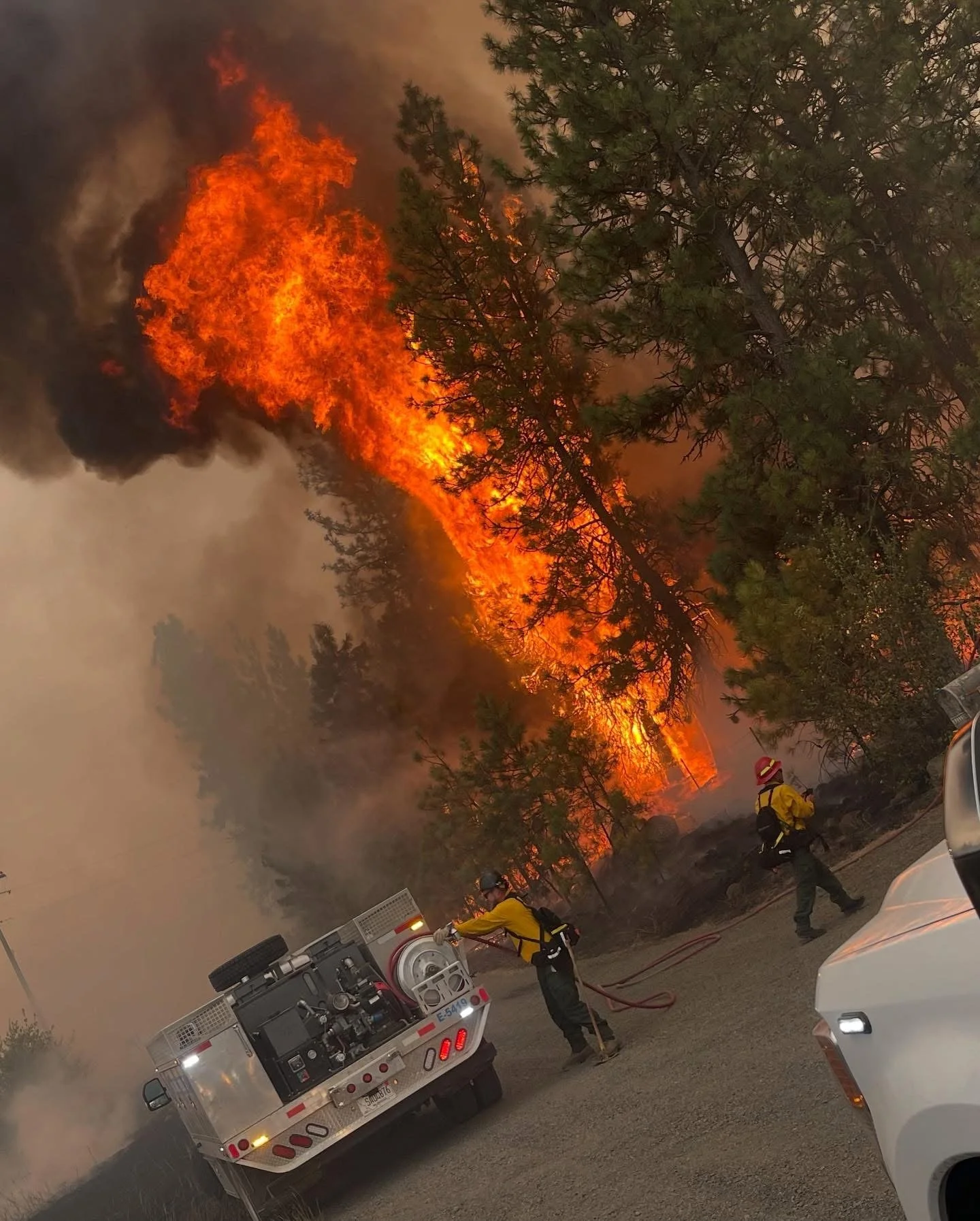 Firefighters are battling a wildfire in a forested area, with large flames and thick smoke. Two firefighters in protective gear are operating a fire hose connected to a fire truck, while other firefighters are nearby. The scene appears intense and ur
