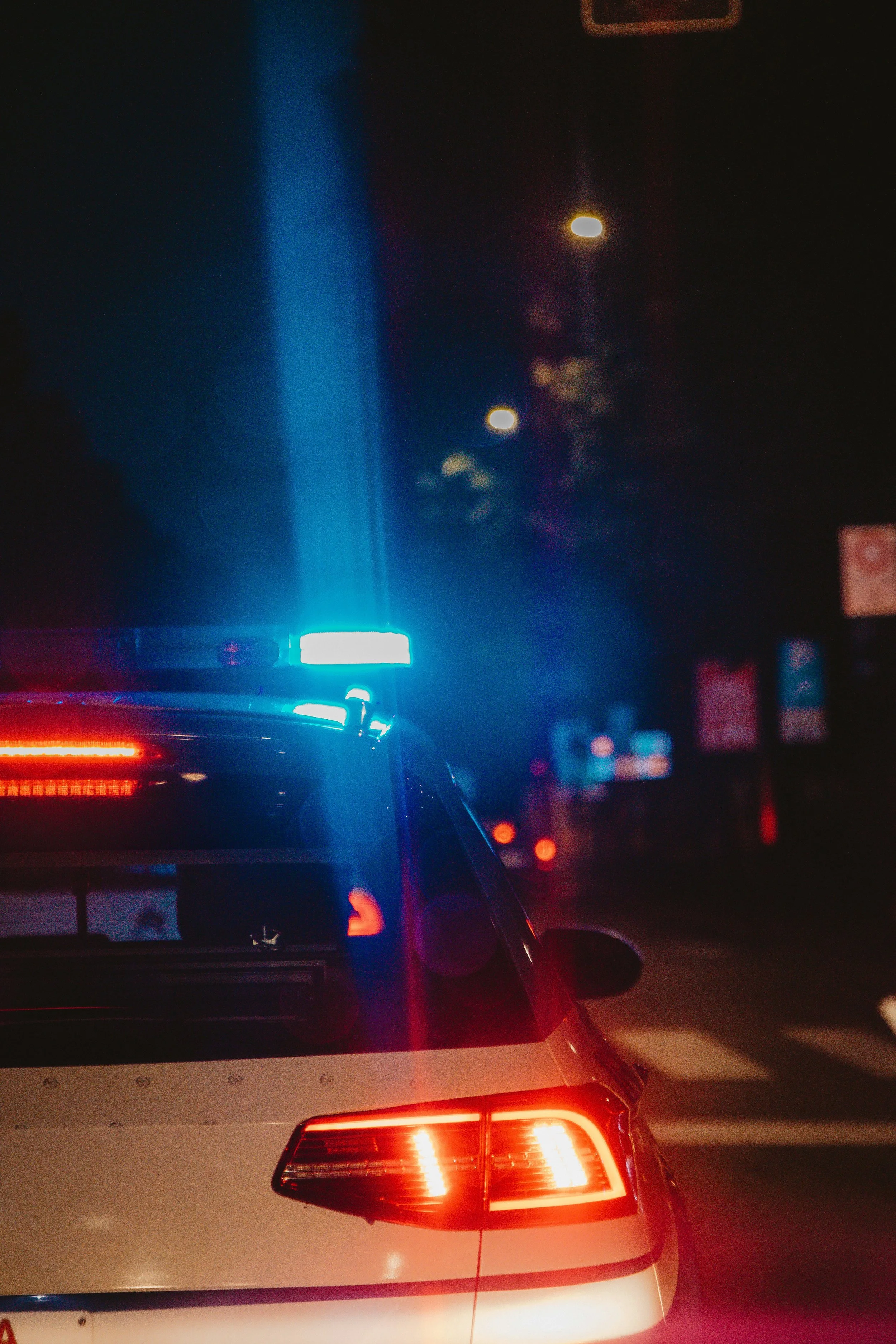 The photo captures the rear view of a white police car with flashing red and blue lights, stopped in traffic at night. The tail lights and part of the traffic lights are visible, with blurred streetlights and signs in the background.
