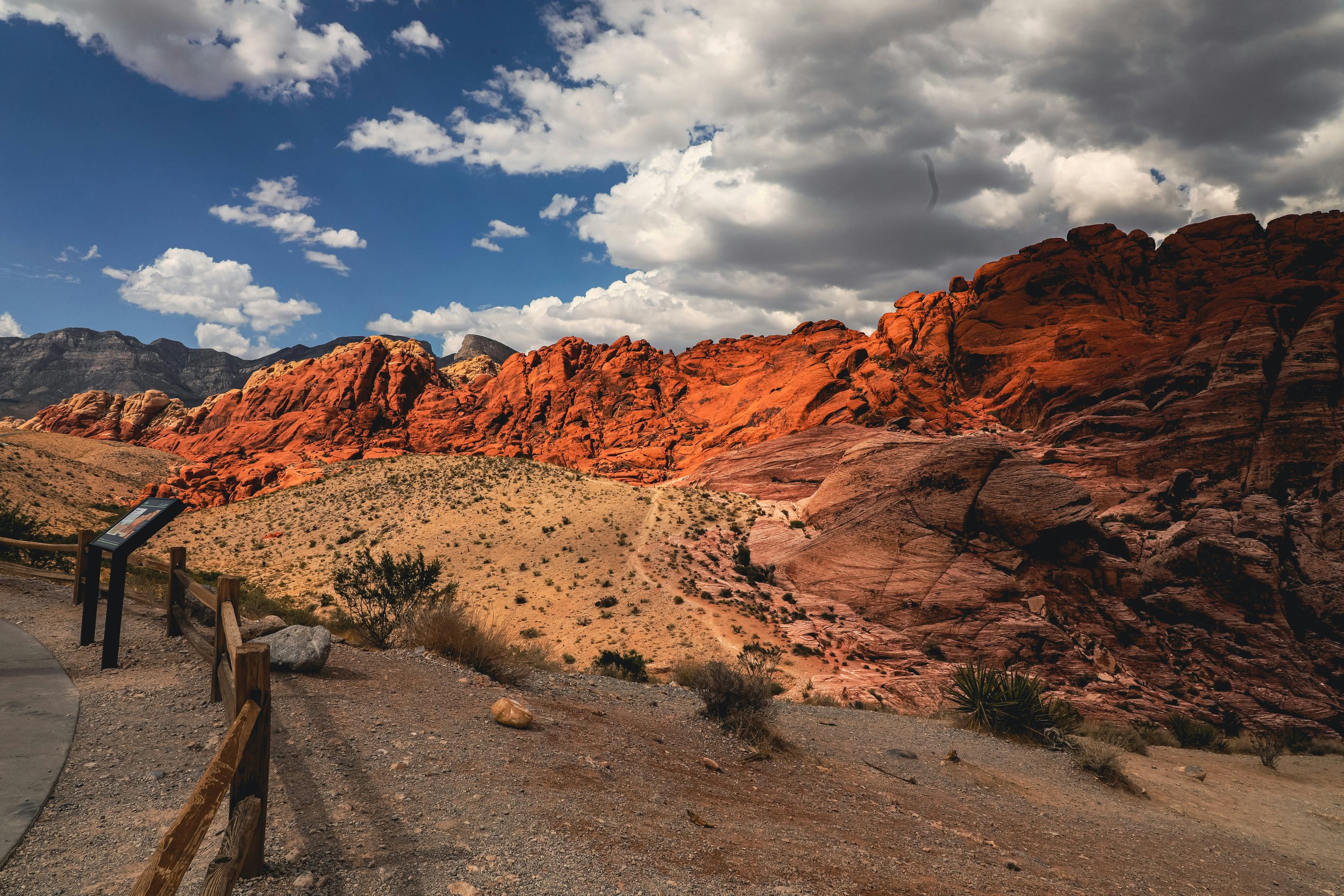 Red rock formations in a desert landscape under a partly cloudy sky, with a gravel path and wooden fence in the foreground.