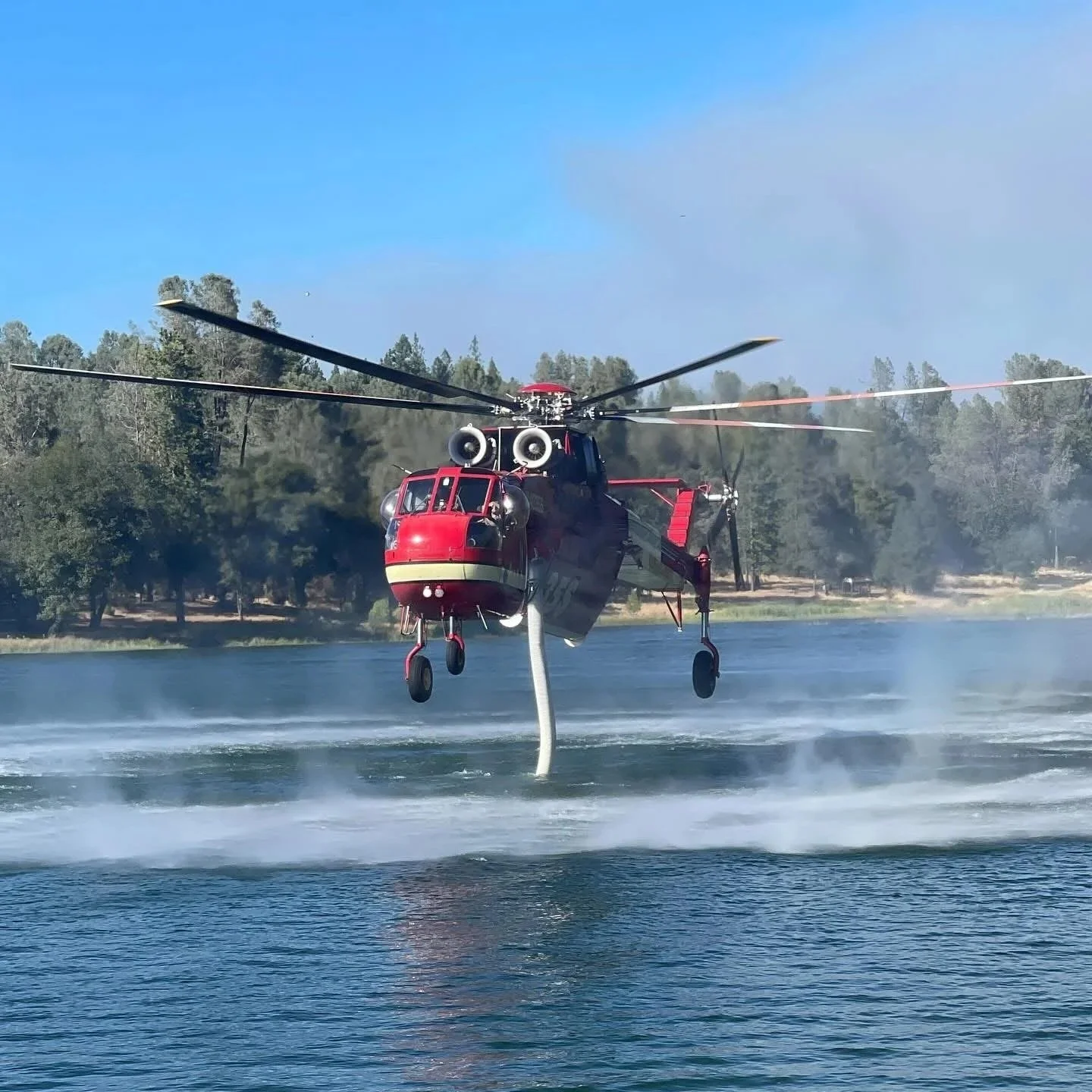 A firefighting helicopter releasing water over a river with trees in the background on a clear day.