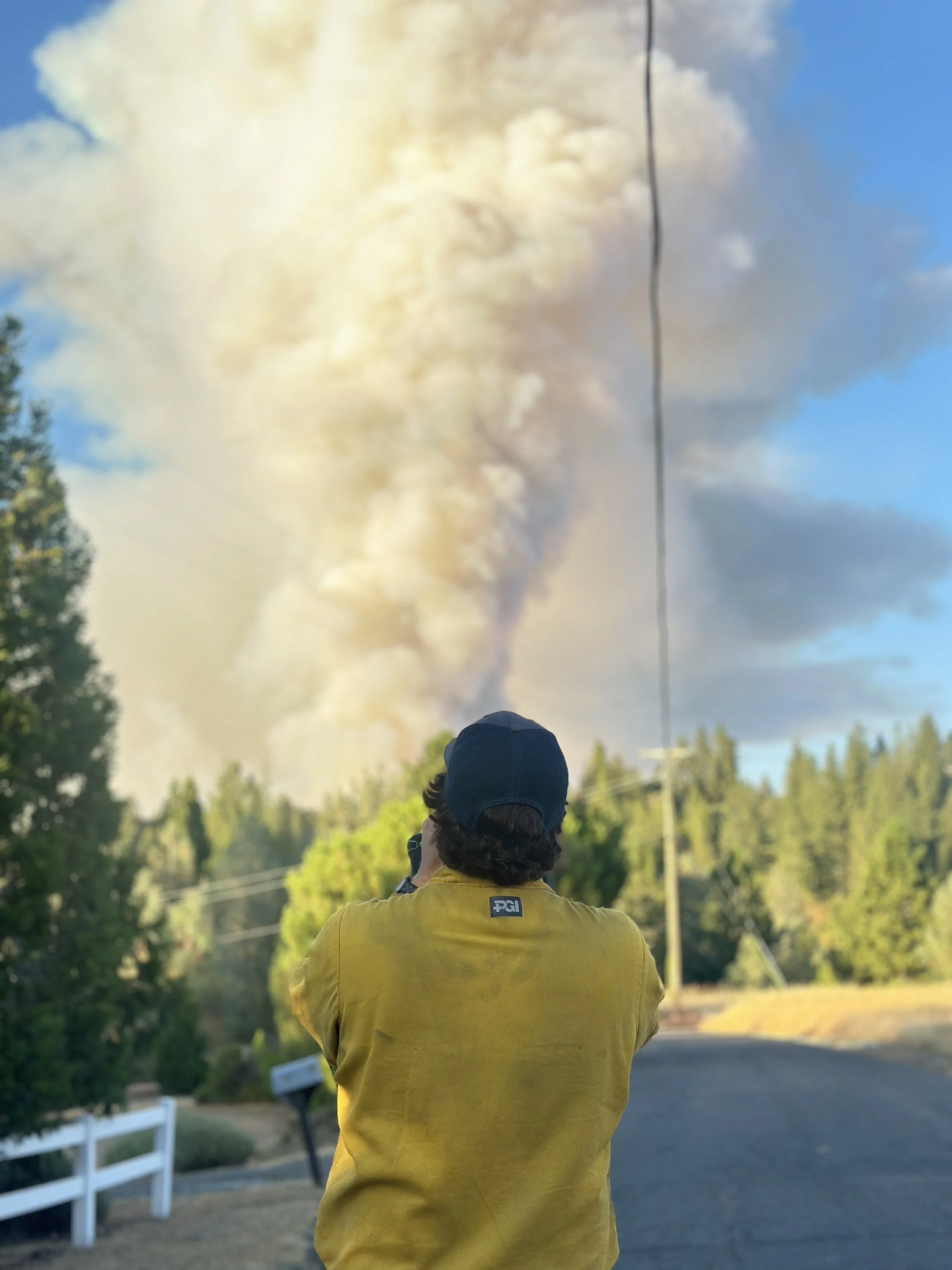 A person with curly hair wearing a yellow jacket and a dark cap is taking a photo with a camera, standing on a road with trees and a white fence, amid a scene of smoke or dust in the sky.