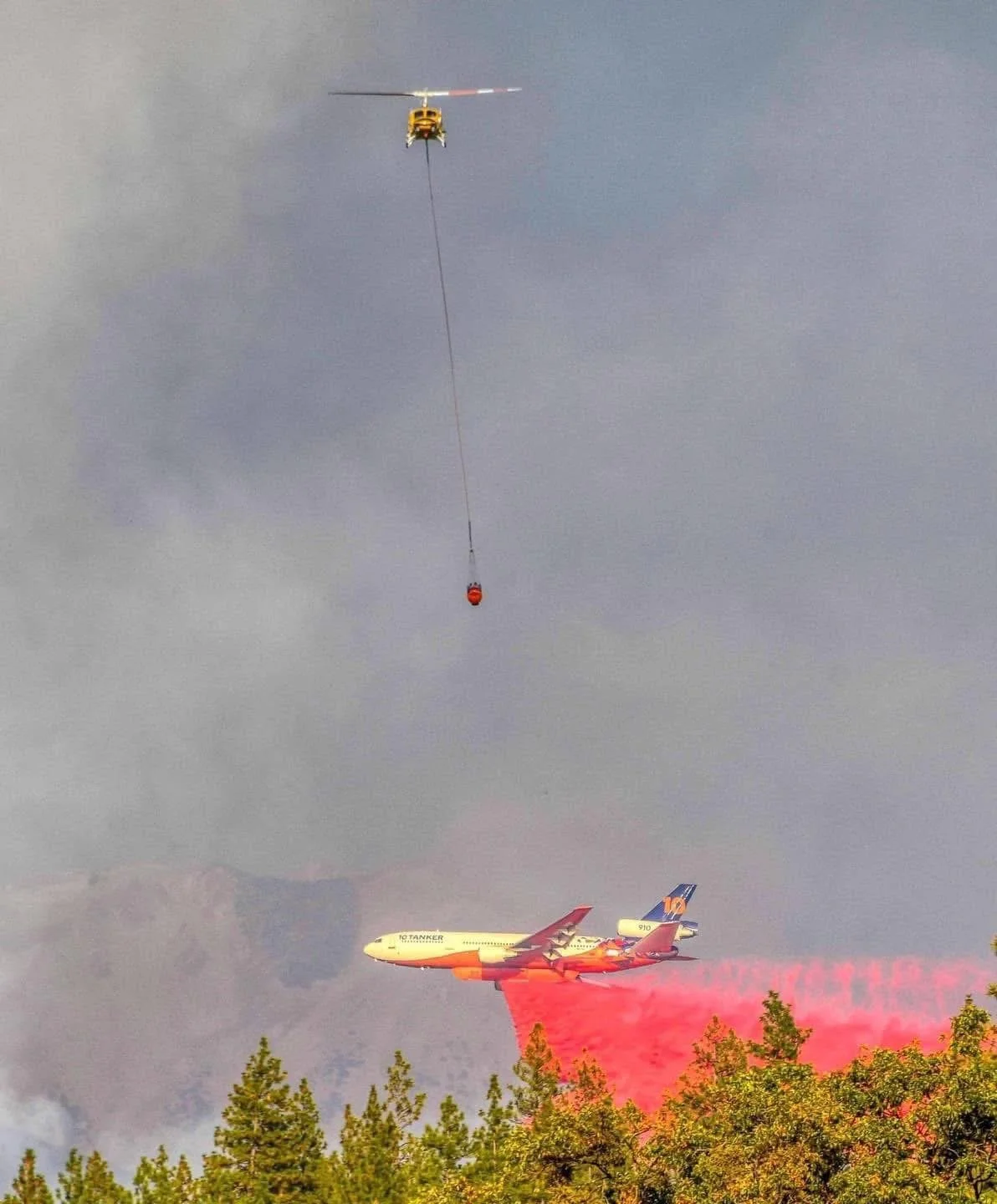 An airplane releasing red fire retardant over a forest fire, with a helicopter above carrying a bucket for water or fire retardant.