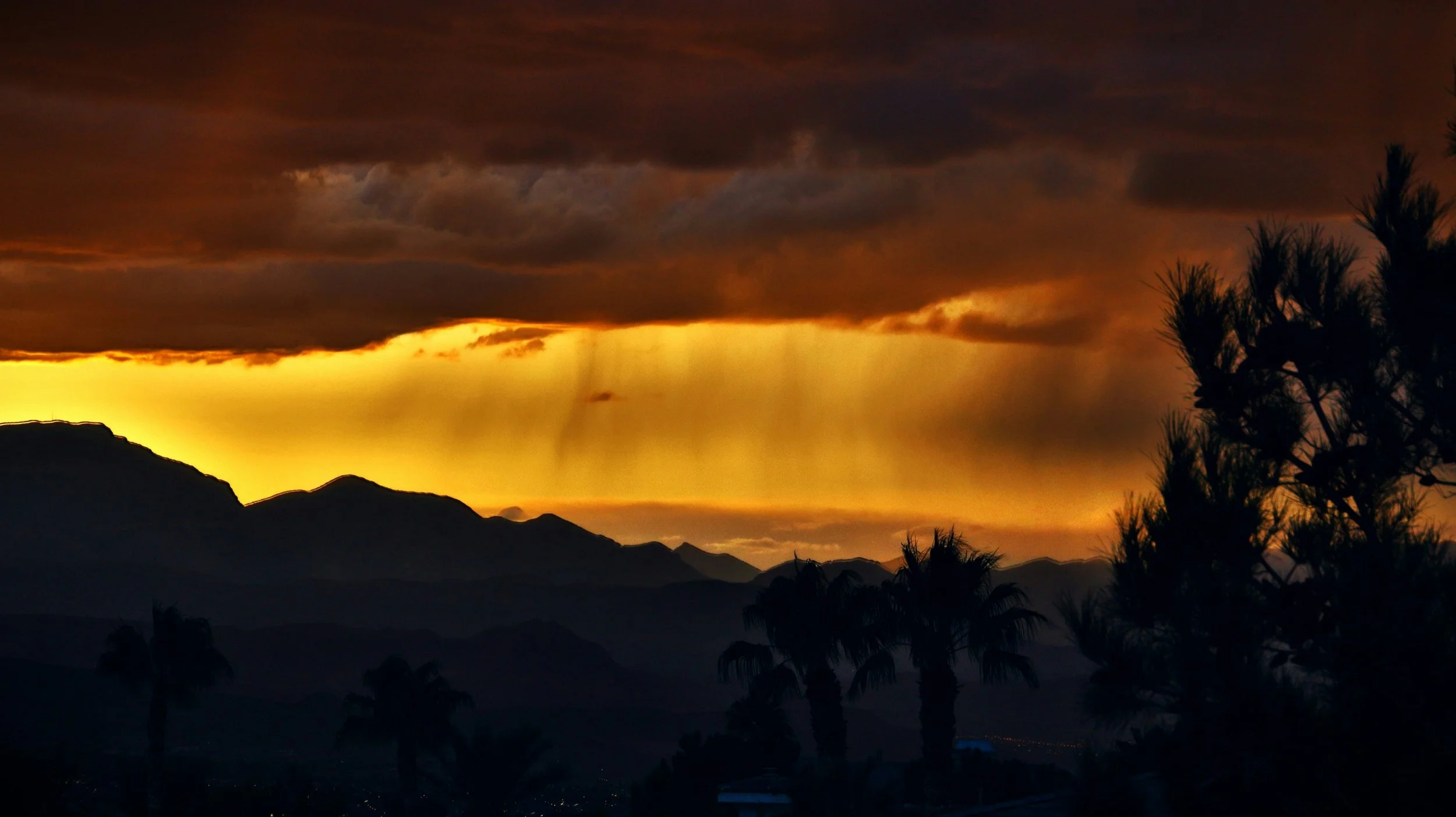 Sunset over mountains with dark clouds and silhouettes of trees in the foreground.