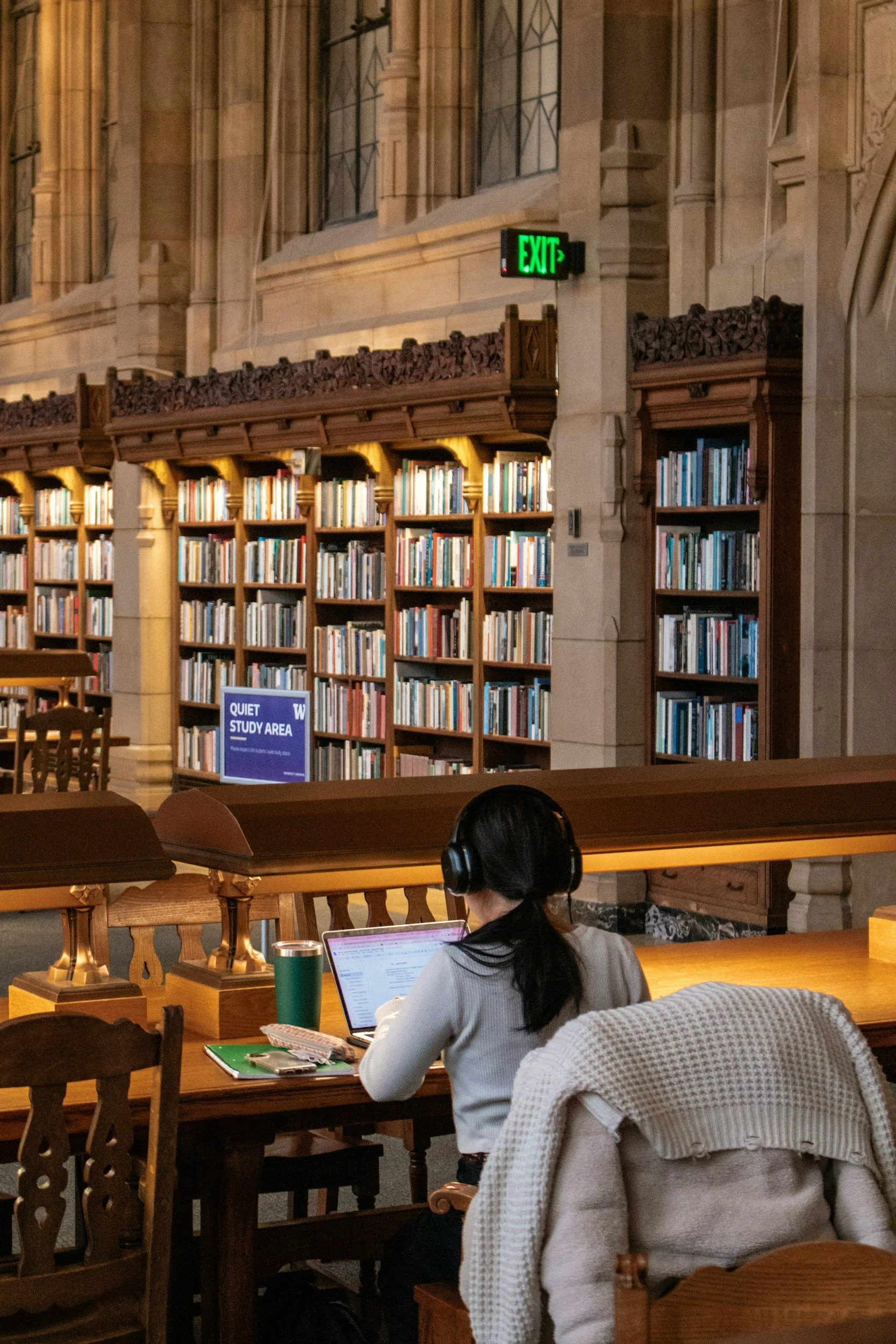 A woman sitting at a wooden table in a library, working on a laptop. She is wearing headphones and a white sweater, with a coat hanging on the chair. There are bookshelves and an 'EXIT' sign in the background.