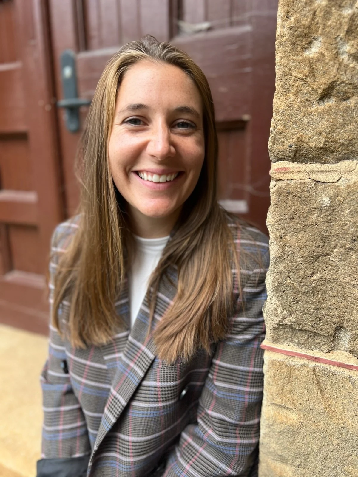 A smiling woman with long light brown hair, wearing a plaid blazer, standing next to a stone wall with a wooden door in the background.