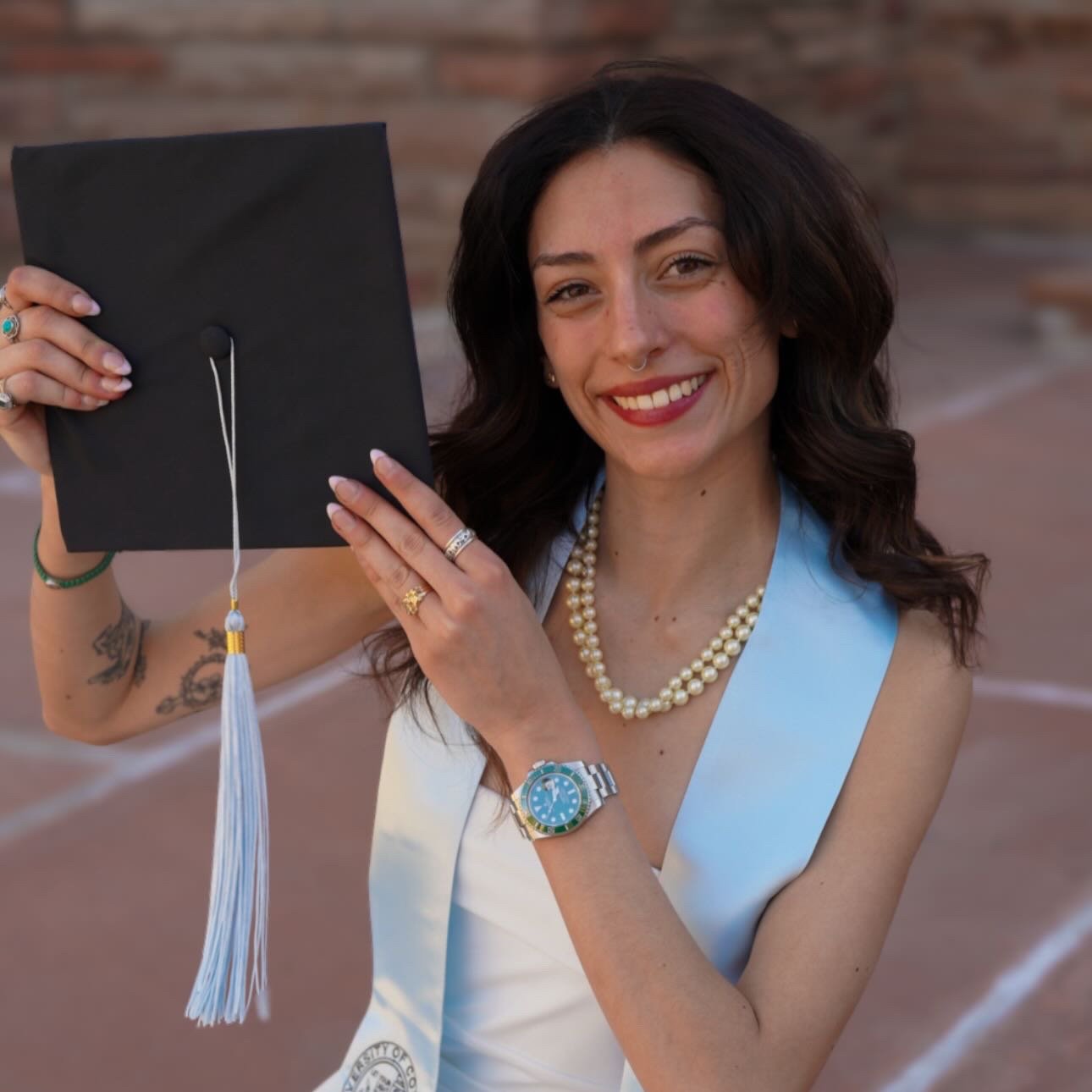 A woman in a light blue dress holding a graduation cap with a tassel, smiling, wearing a pearl necklace, a watch, and rings, standing on a track field.