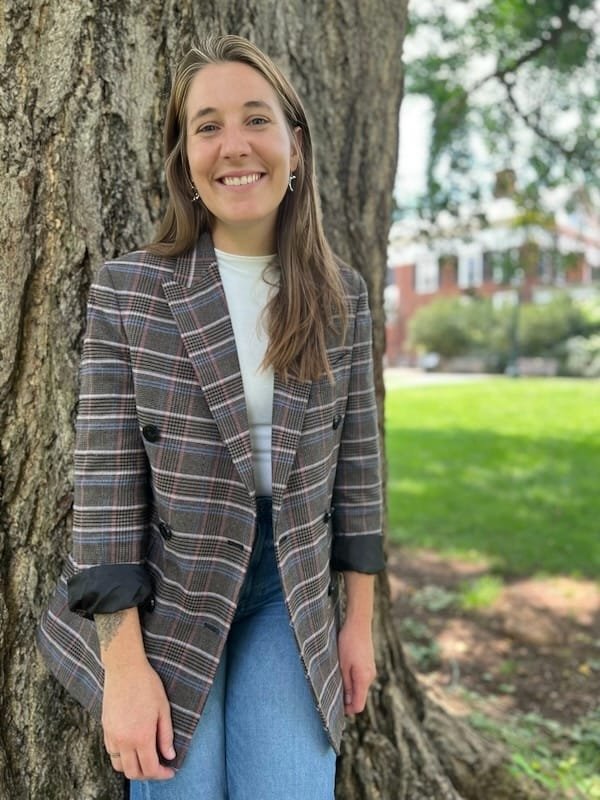 A woman with long brown hair, smiling, standing outdoors against a large tree. She is wearing a plaid blazer over a white shirt and blue jeans.