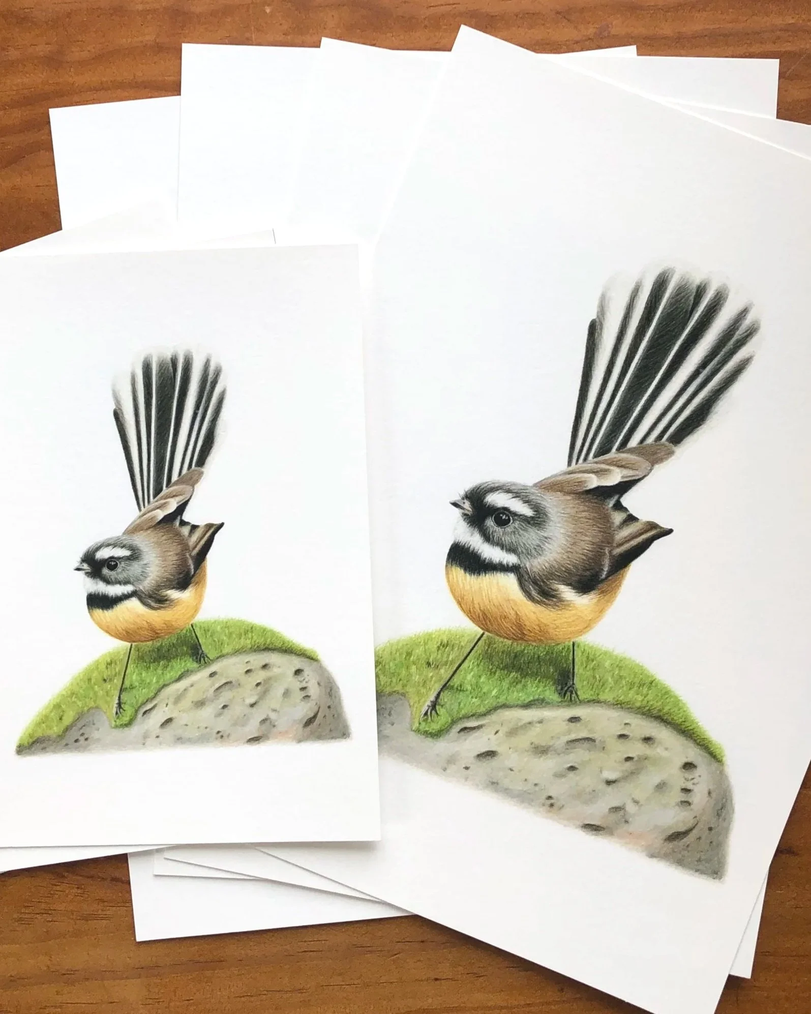 Multiple sheets of paper with a drawing of a bird with a birdhouse on green grass, on a wooden surface.