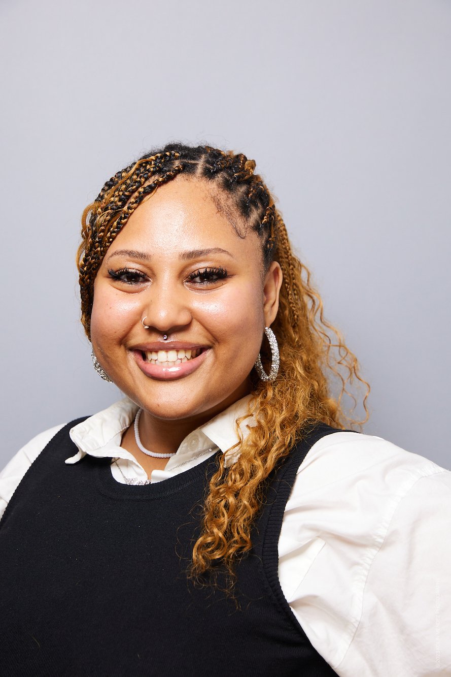 Smiling woman with blonde braids, hoop earrings, a necklace, and a nose ring, wearing a white shirt and black vest, against a neutral background.