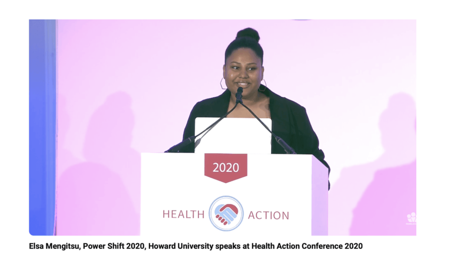 A woman speaking at a conference, standing behind a podium with a sign displaying the year 2020 and a logo with two hands. The background is pink, and the woman is wearing a dark blazer with hoop earrings.