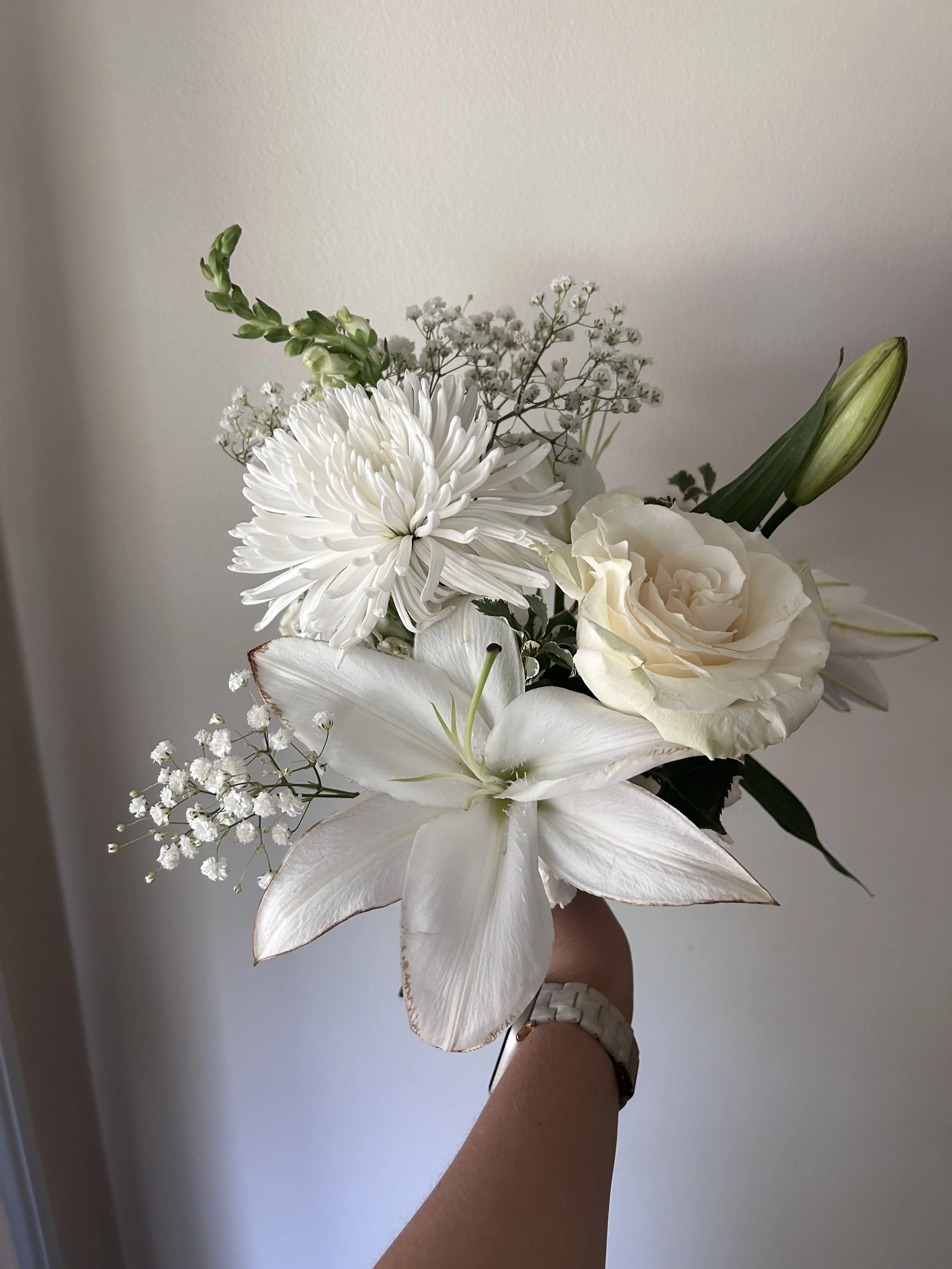 A hand holding a bouquet of white flowers, including a large white lily, a white rose, a white chrysanthemum, and small white filler flowers, against a plain wall background.