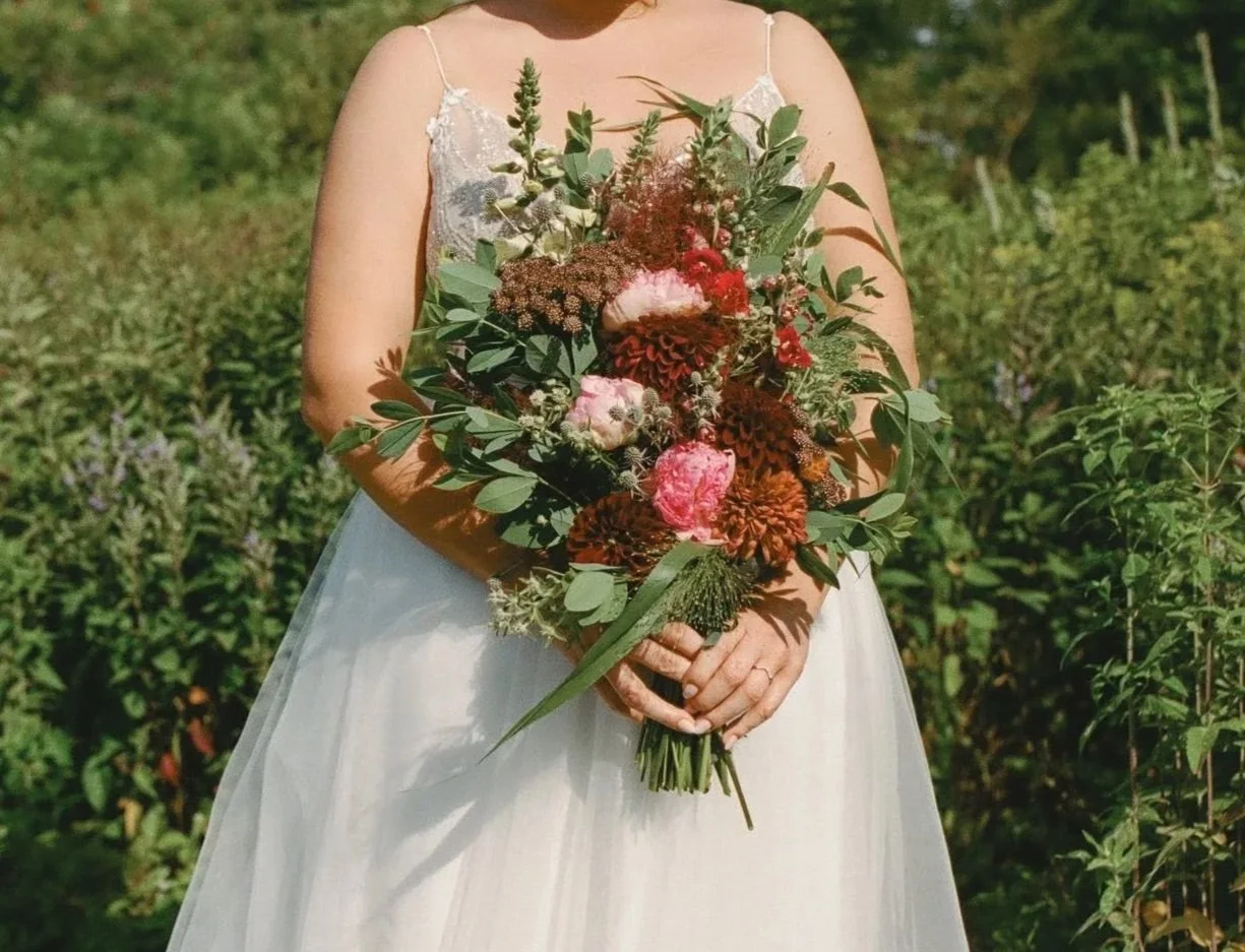 A bride in a white wedding dress holding a large bouquet of mixed flowers and greenery outdoors in a field.